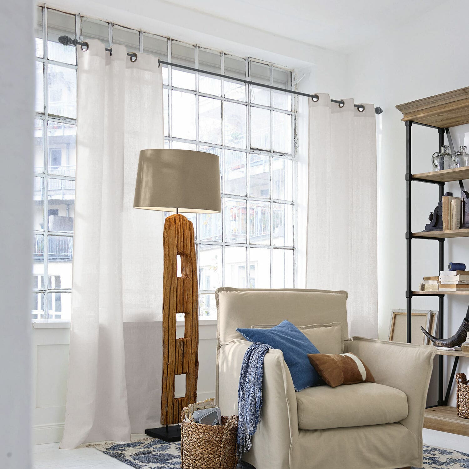 Gardine Salinas Stehlampe mit Holzfuß und beigem Lampenschirm in hellem Wohnzimmer mit Sessel und Regal