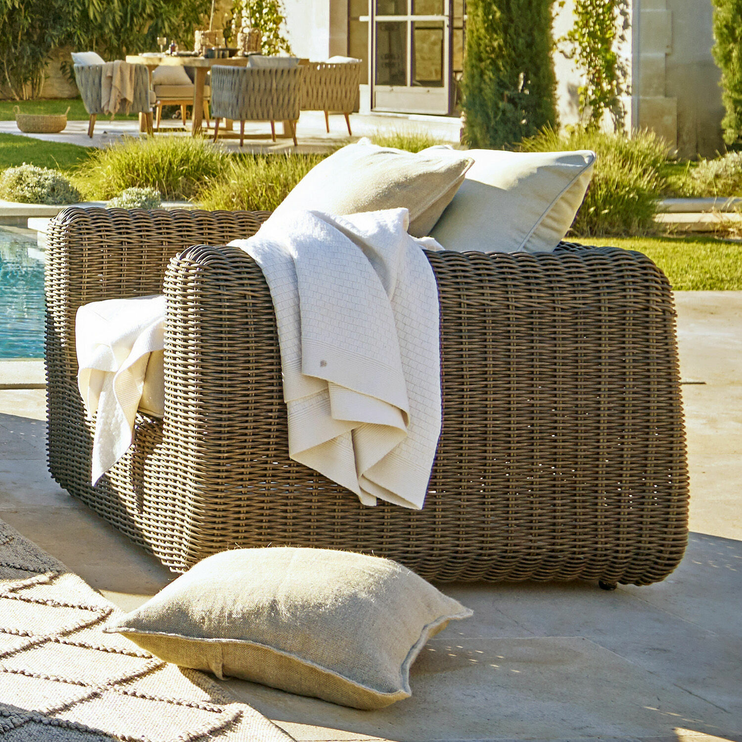 Gartenfauteuil aus braunem Geflecht mit hellen Kissen und Decke am Pool