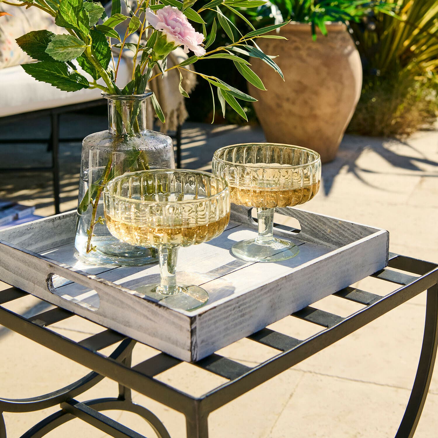 Zwei transparente Champagnergläser auf weißem Holztablett auf einem Metall-Gartentisch im Sonnenlicht