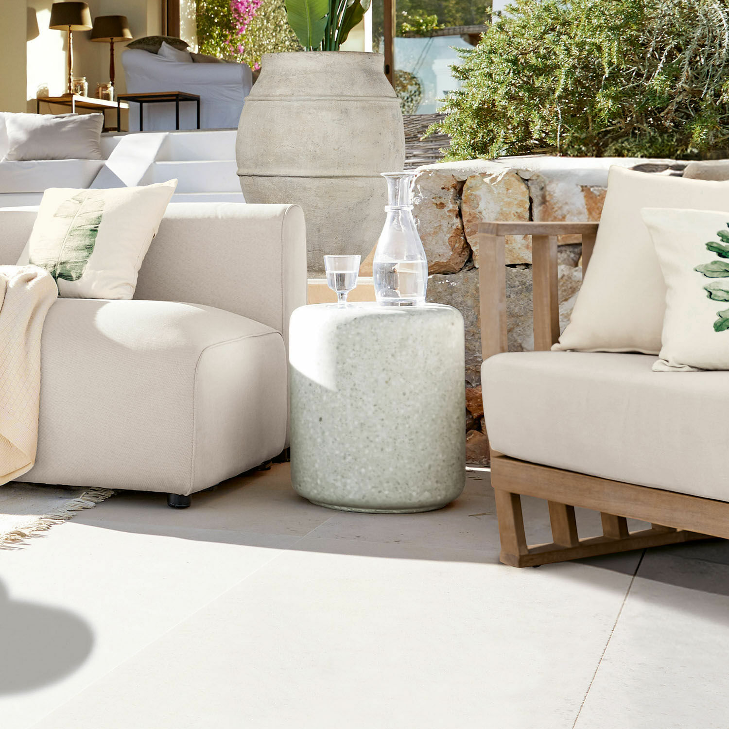 Runder Beistelltisch aus hellem Beton auf Terrasse mit beigen Sofas und dekorativer Wasserkaraffe