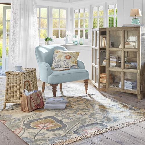 Hellblauer Sessel mit gemustertem Kissen in hellem Wohnzimmer mit Teppich und rustikalem Holzschrank