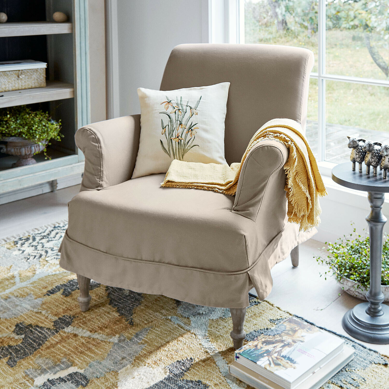 Sessel Everleighs Beiger Polstersessel aus Stoff mit floralem Kissen und gelbem Plaid in hellem Wohnzimmer mit Teppich