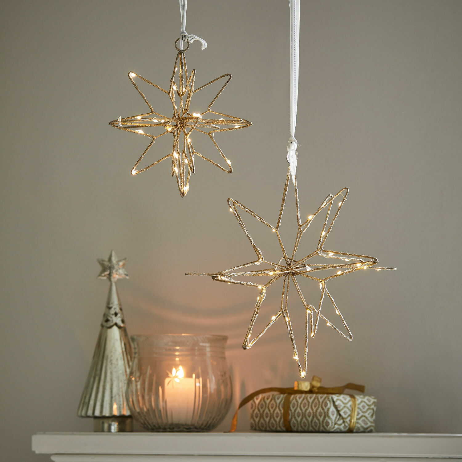 LED-Stern 2er Set Pérole Goldene LED-Stern-Dekorationen hängen über Kerze und Geschenk auf weißem Regal