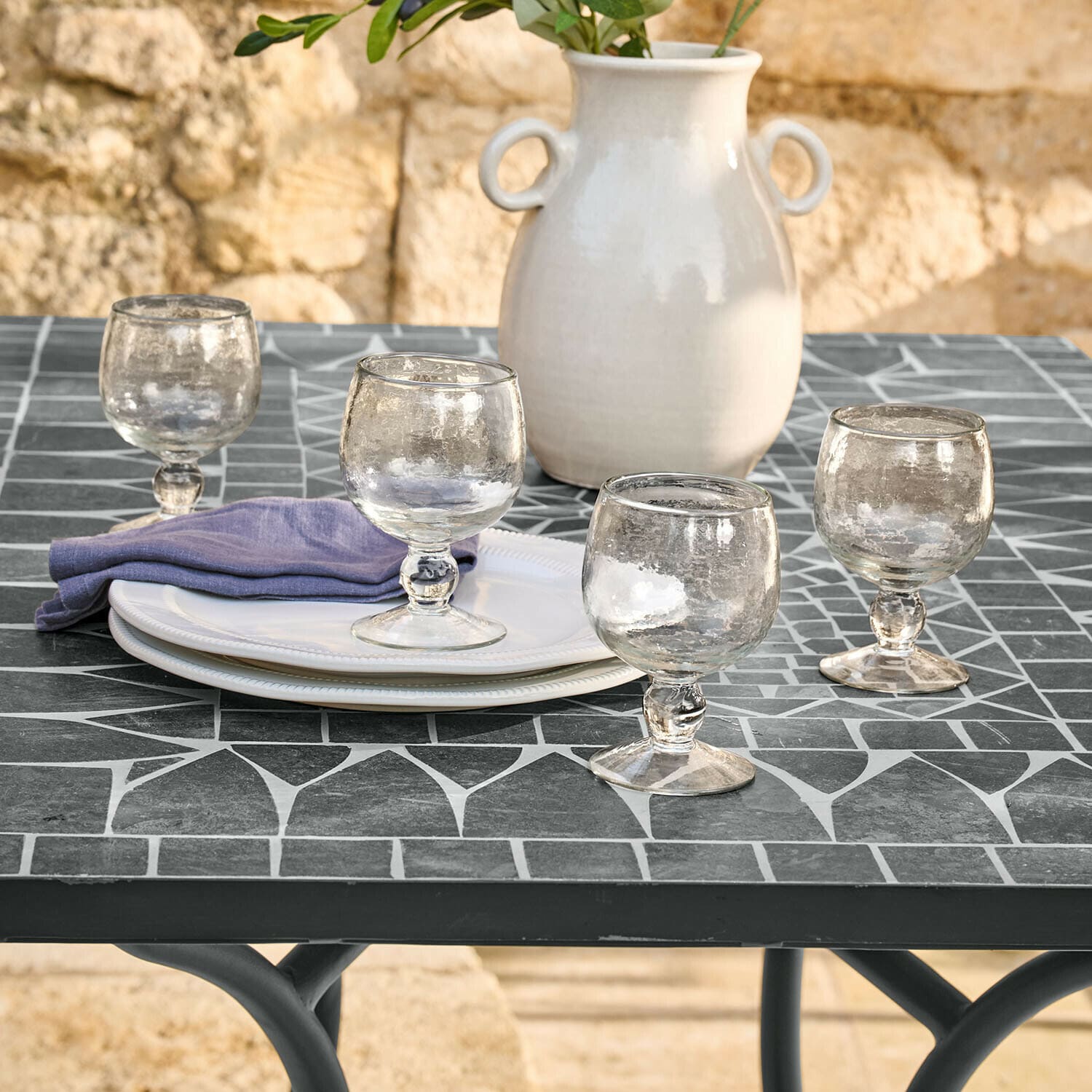 Gläser aus Glas auf grauem Mosaiktisch mit weißen Tellern und weißer Keramikvase mit grünen Blättern