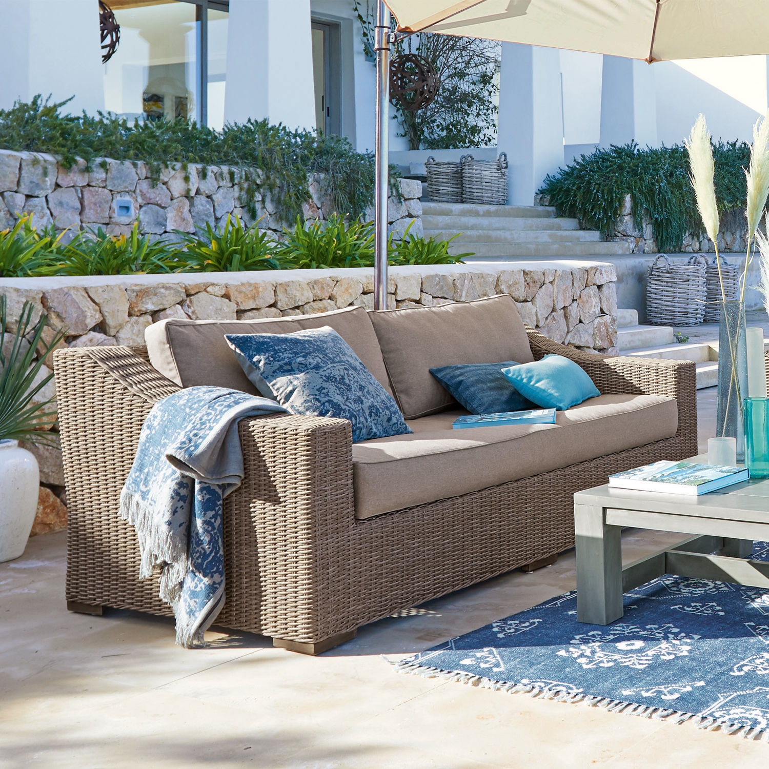 Beiges Rattan-Gartensofa mit blauen Kissen und Decke auf sonniger Terrasse