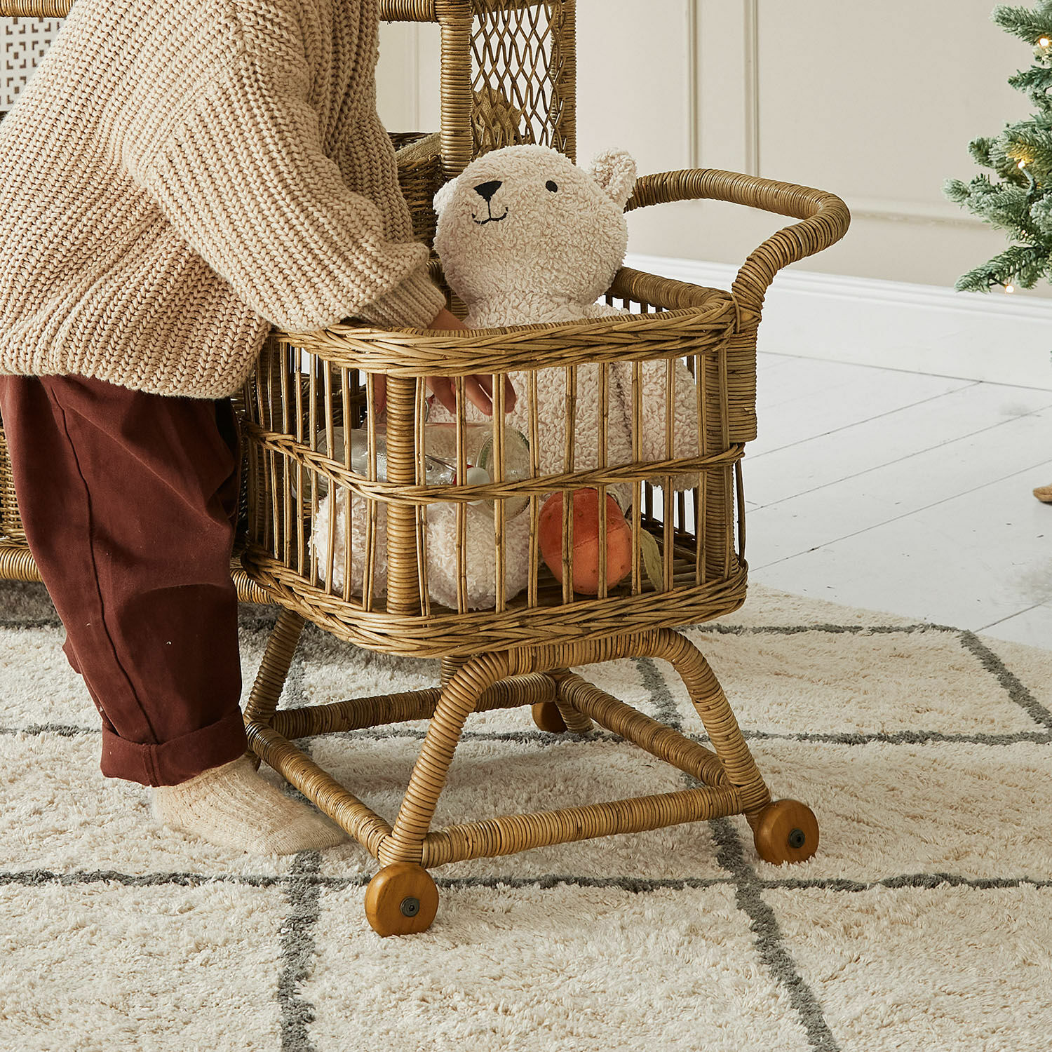 Kind spielt mit einem Rattankinderwagen mit Plüschbär und Spielzeug auf hellem Teppich im Wohnzimmer