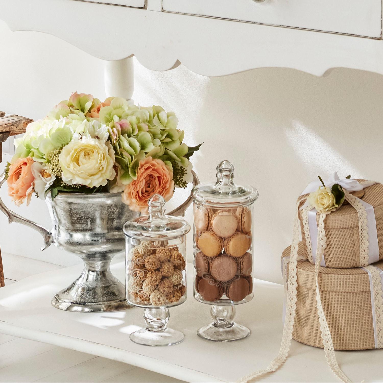 Dekorativer silberner Blumenpokal mit Glasgefäßen voller Macarons und Geschenken auf weißem Tisch