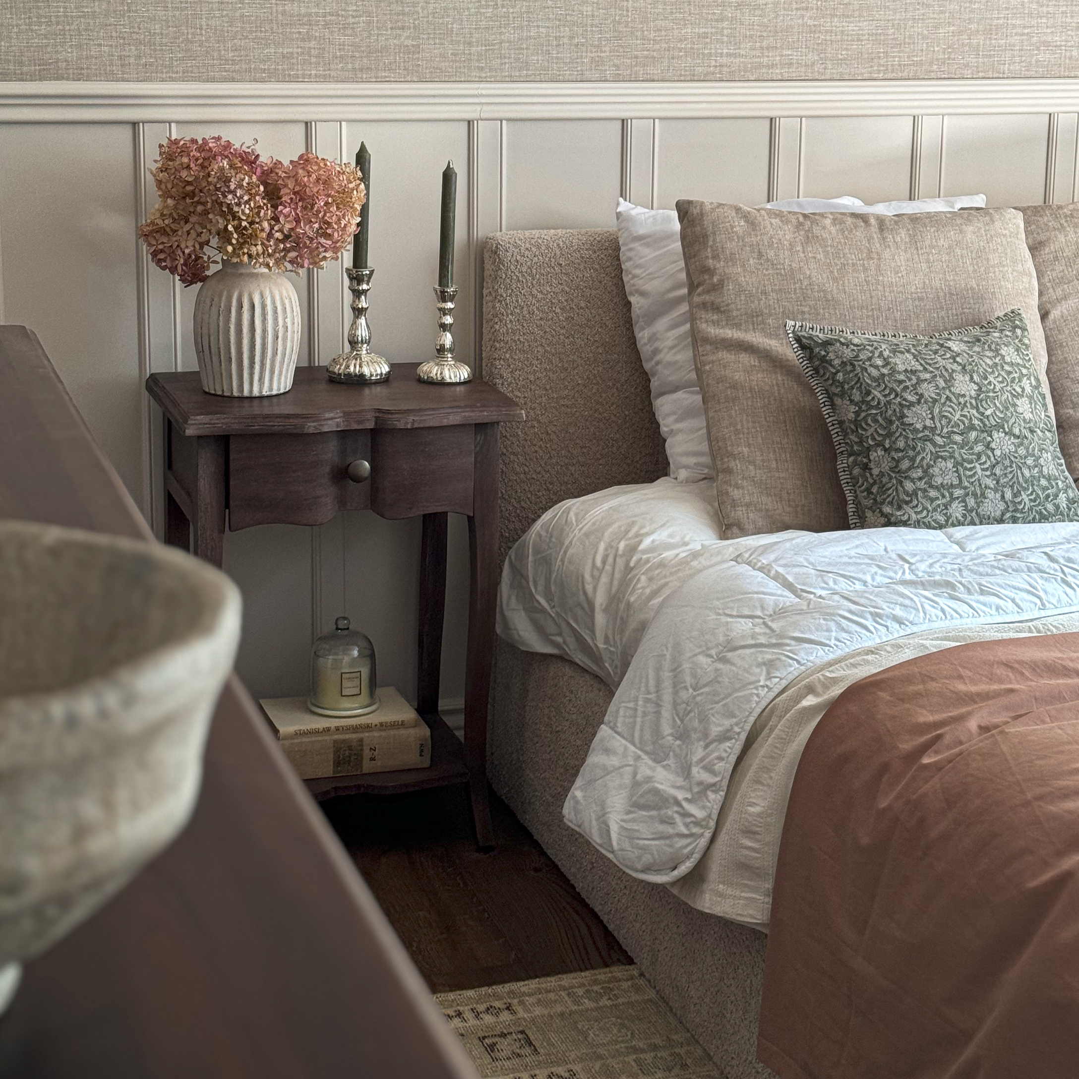 Gemütliches Schlafzimmer mit beiger Bettwäsche, dunklem Holznachttisch und Vase mit getrockneten Blumen