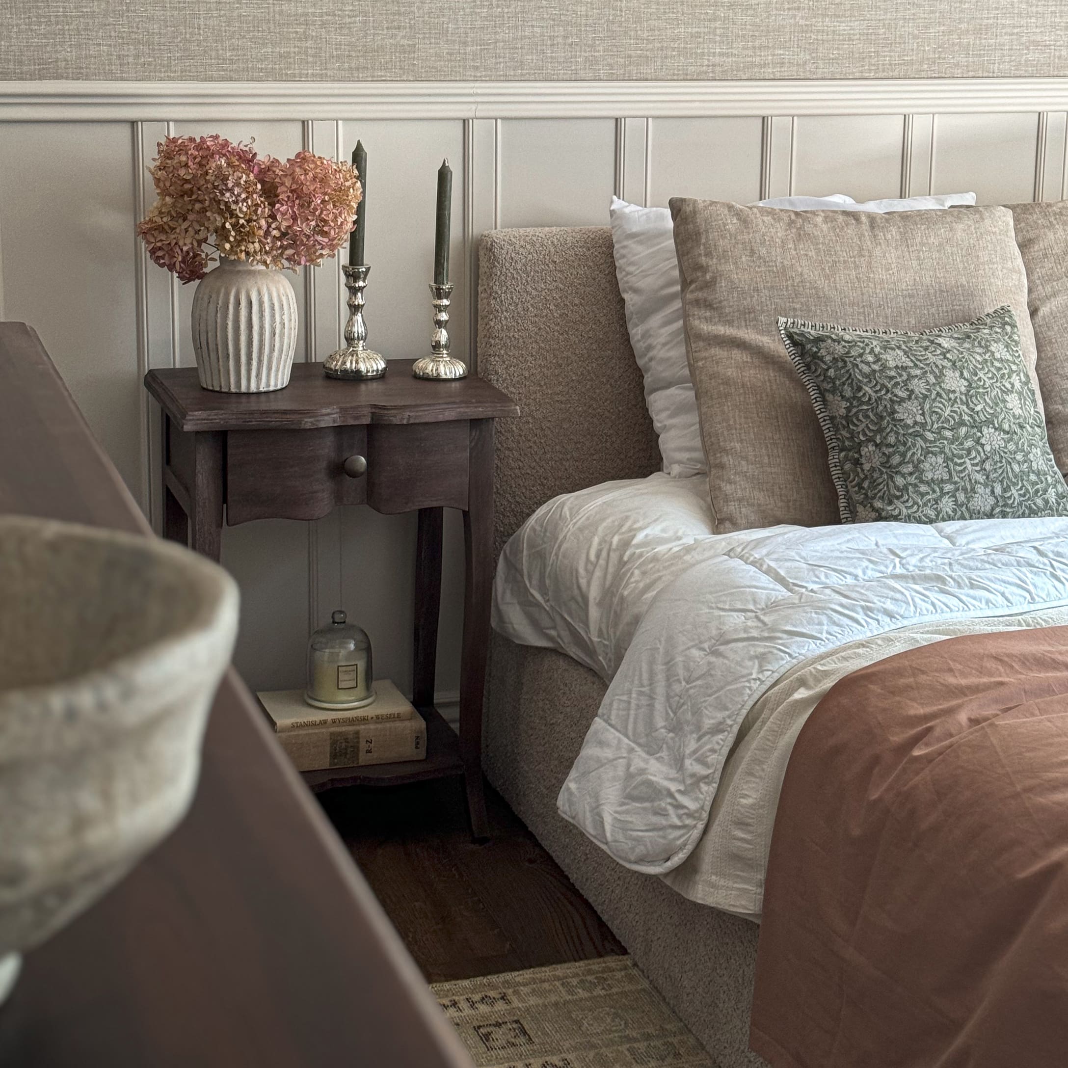 Gemütliches Schlafzimmer mit beiger Bettwäsche, dunklem Holznachttisch und Vase mit getrockneten Blumen