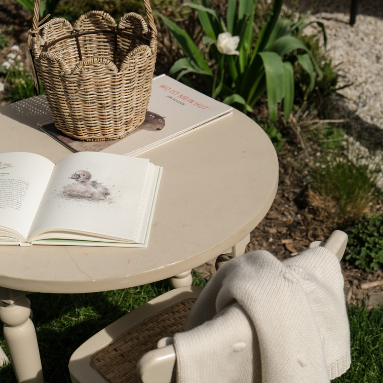 Runder beiger Gartentisch mit Weidenkorb und Büchern auf dem Rasen bei Sonnenschein