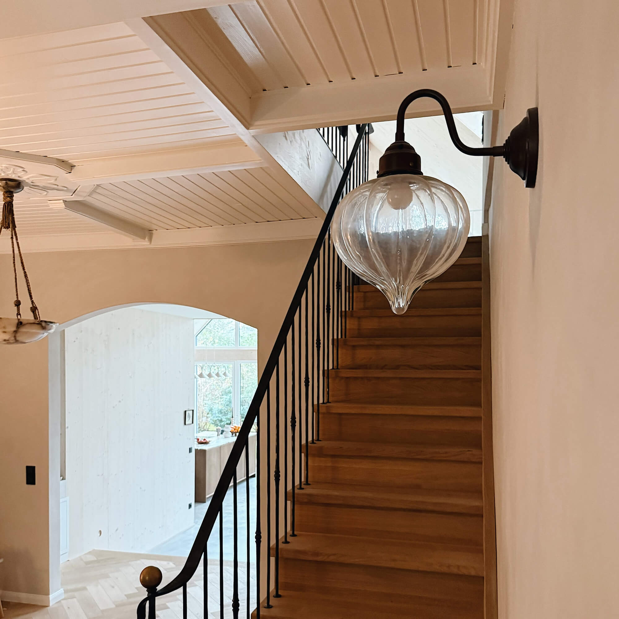 Glas-Wandleuchte mit schwarzem Metallarm über Holztreppe in hellem Wohnraum