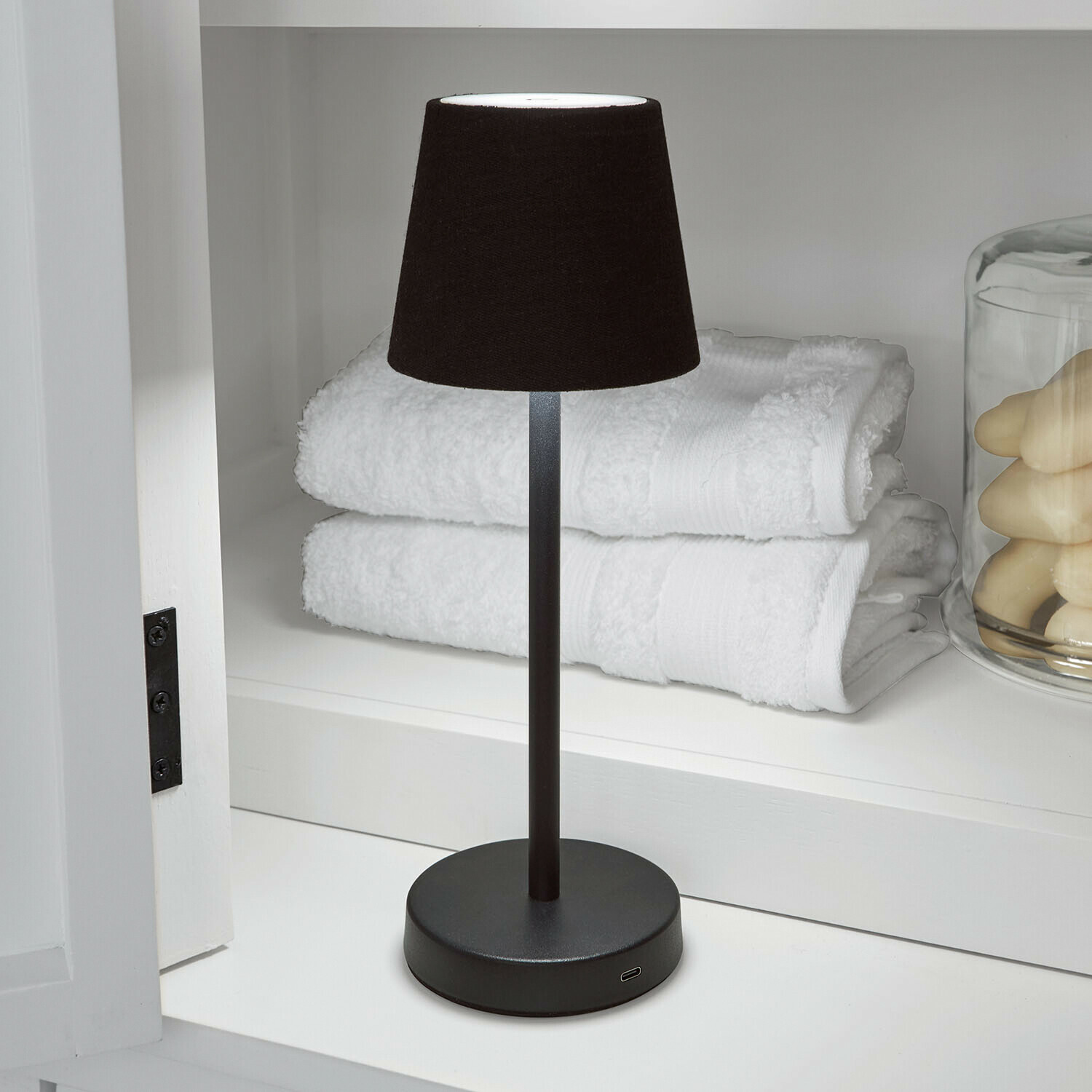 Wiederaufladbare Tischlampe Sylvainie Schwarze Tischlampe mit Stoffschirm auf Metallfuß neben weißen Handtüchern