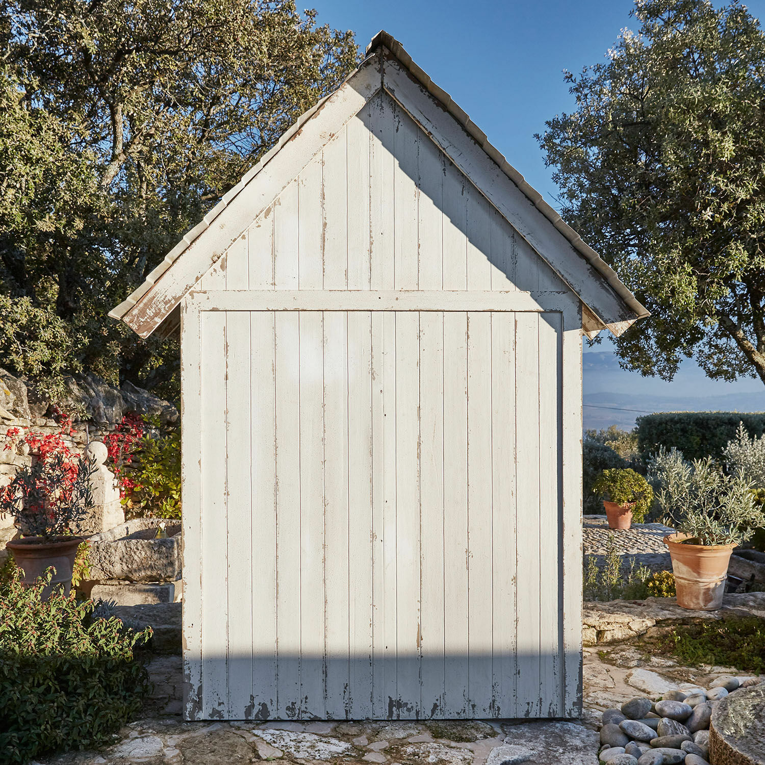 Weißes Holz-Gartenhaus mit verwitterter Oberfläche auf steinigem Boden umgeben von Pflanzen