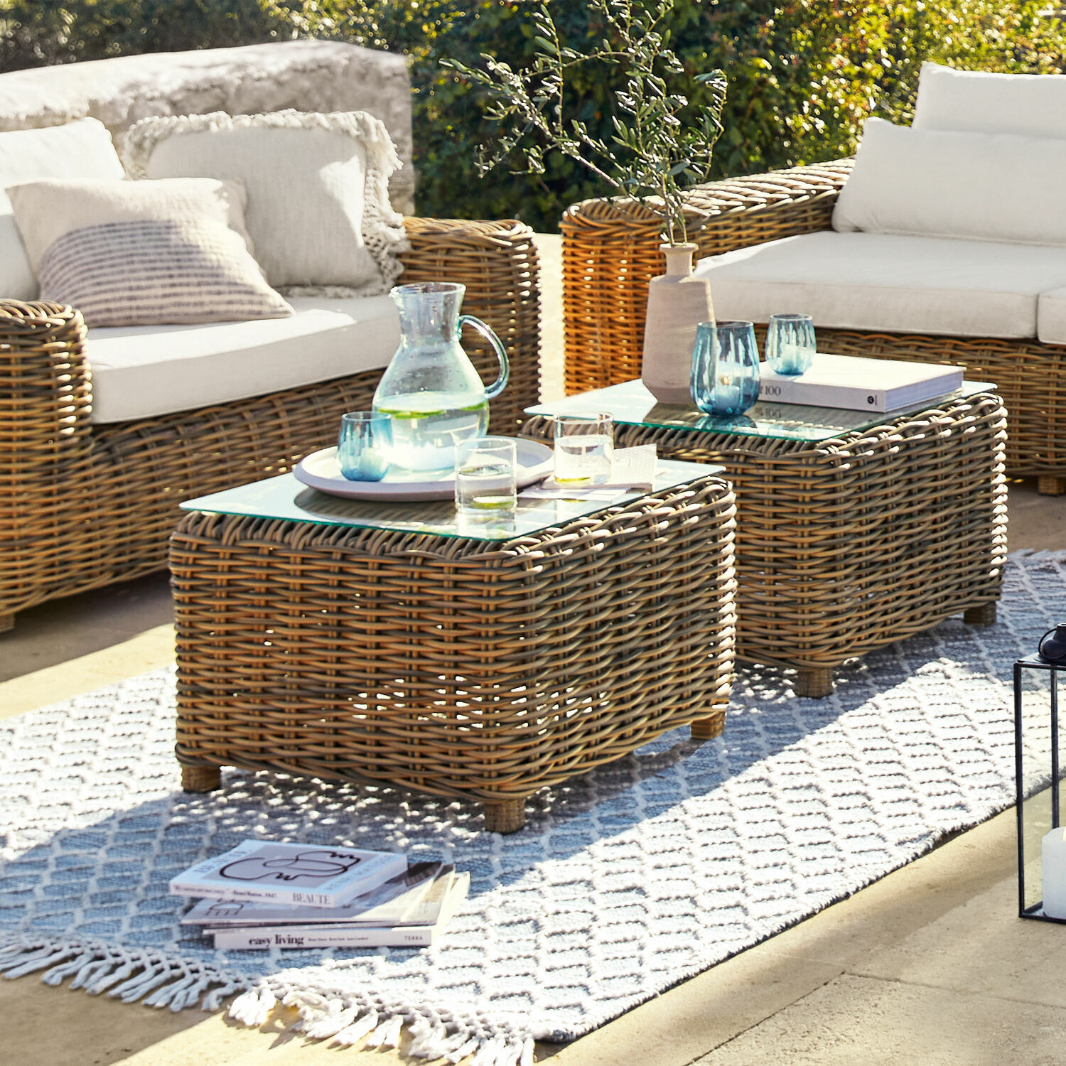 Gartenmöbel-Set aus Rattan mit weißen Kissen und Glastischen auf Terrasse im Sonnenlicht