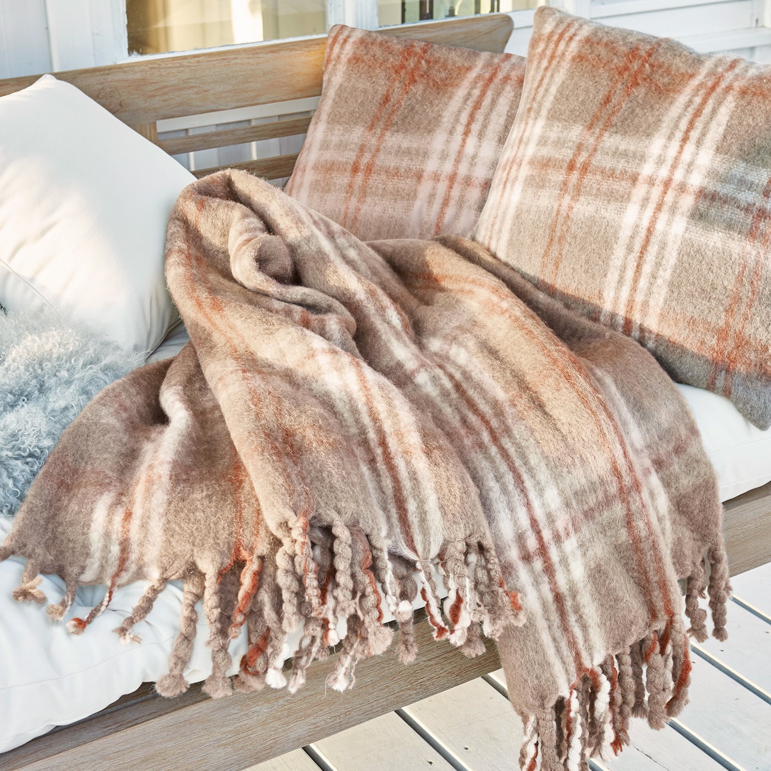Weiche beige Kuscheldecke und kariert gemusterte Kissen mit Fransen auf Holzbank auf Terrasse