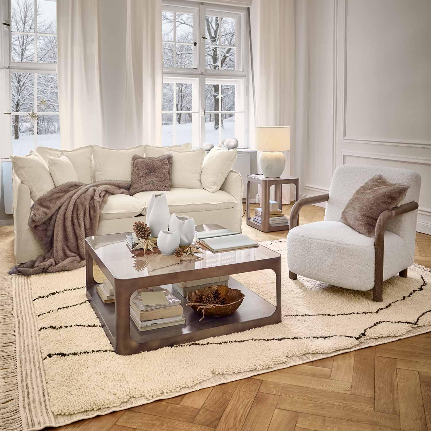 Gemütliches Wohnzimmer mit weißem Sofa, Sessel mit Holzarmlehnen und weichem Teppich im Winterambiente