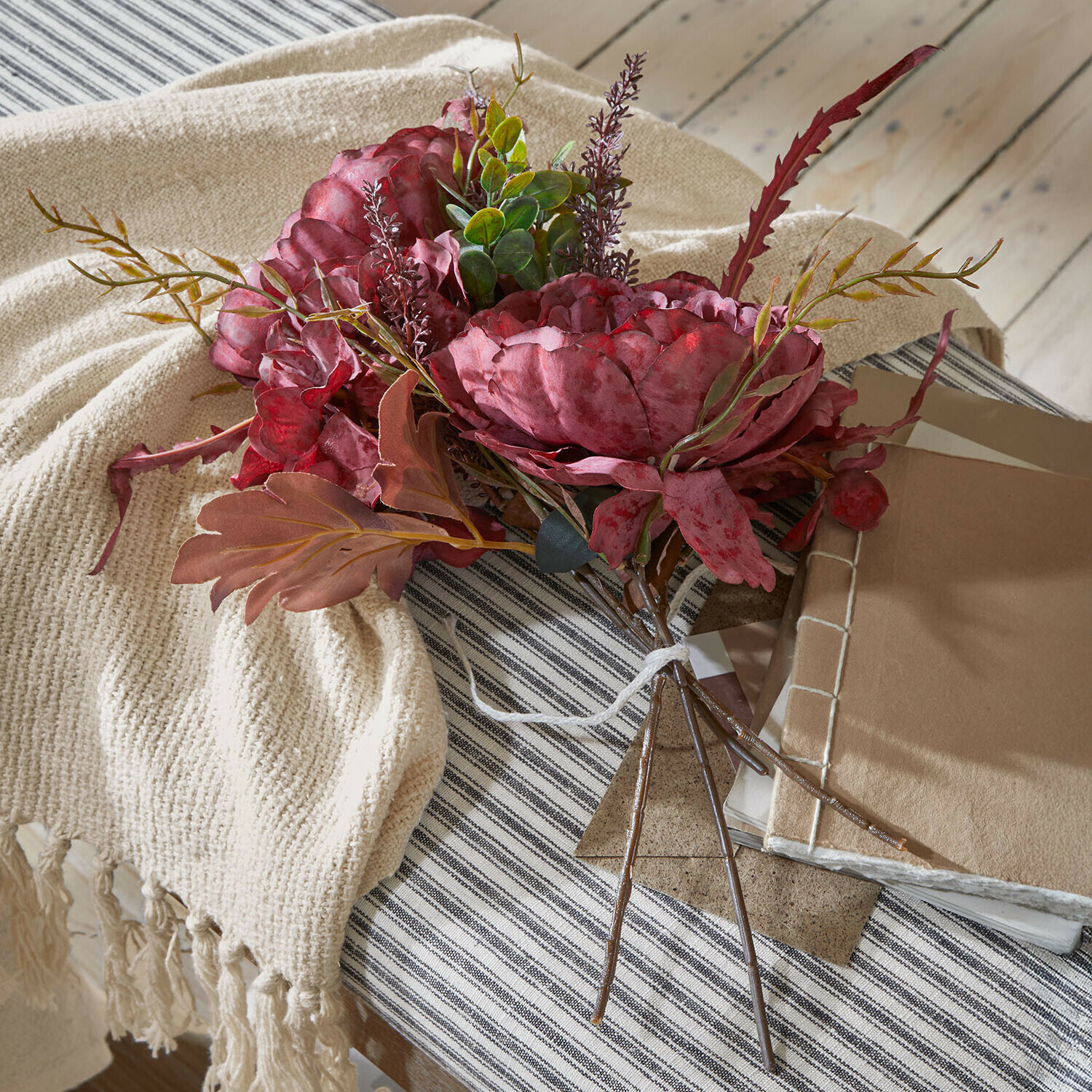 Deko-Blumenstrauß Rivière Ambrée Strauß aus künstlichen roten Blüten mit grünem Laub auf beigem Plaid und gestreiftem Tischtuch