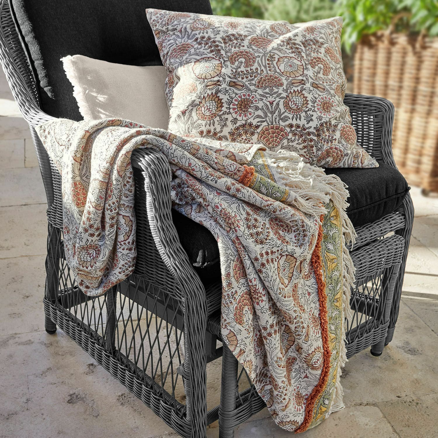 Plaid Romila Baumwoll-Decke und Kissen mit warmem Blumenmuster auf grauem Rattansessel für gemütliche Atmosphäre