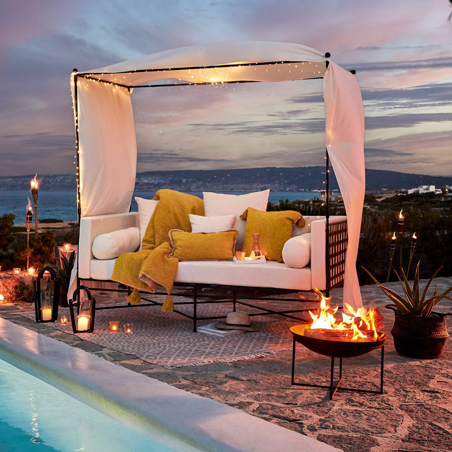 Weiße Metallsofa mit gelben Kissen und Decke am Pool mit Lichterketten und Feuerstelle