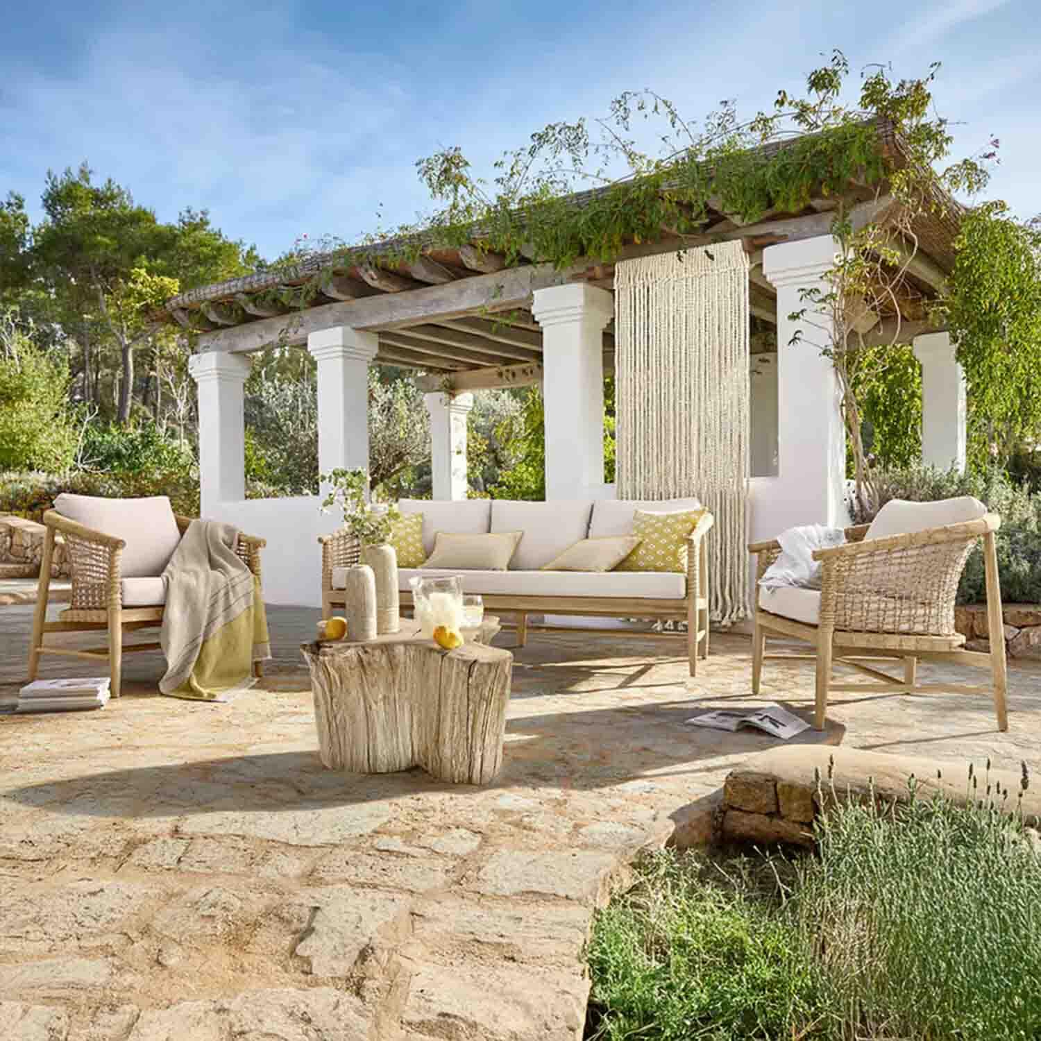 Gartenmöbel-Set aus hellem Holz mit cremefarbenen Polstern auf Steinterrasse unter begrüntem Pavillon