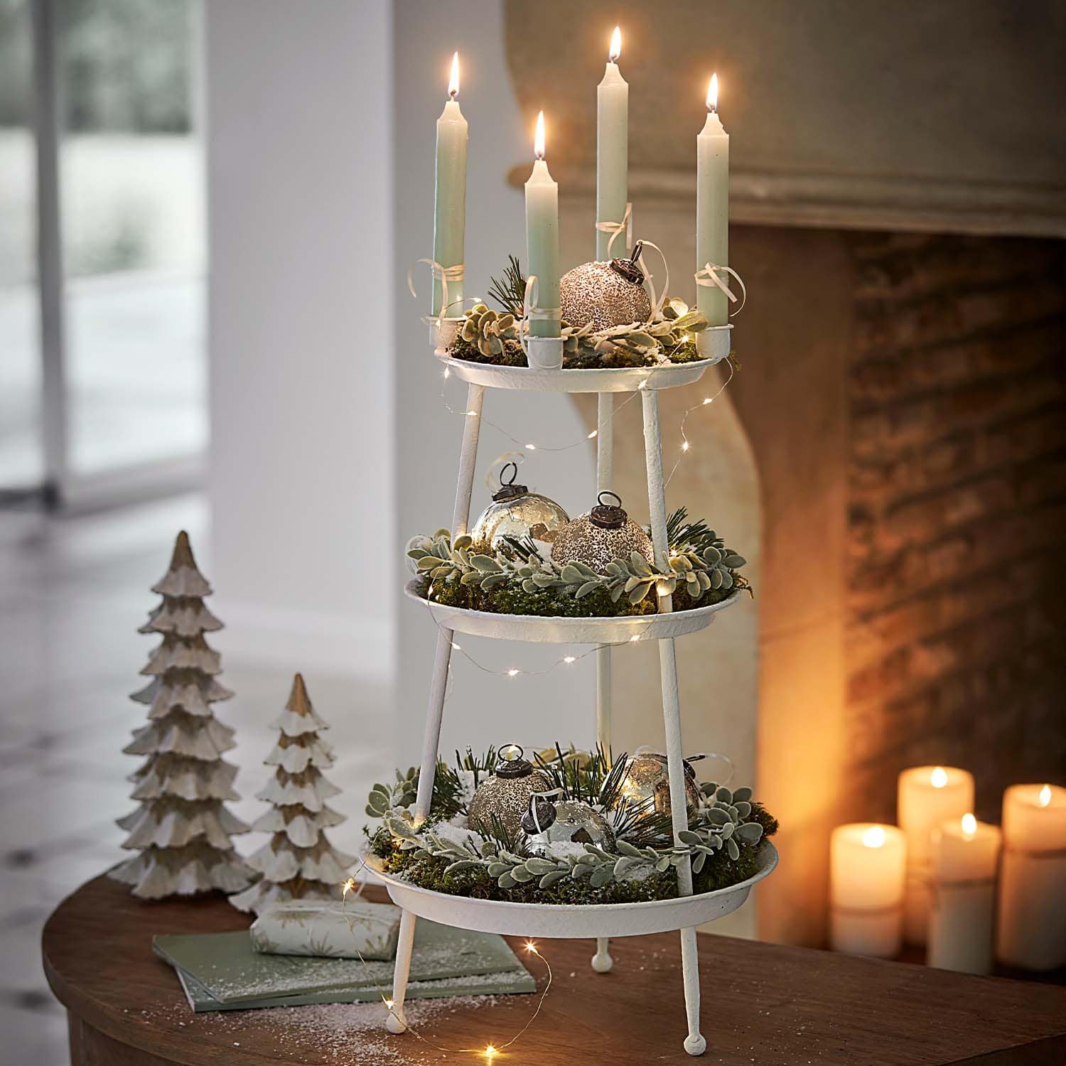 Dreistufige weiße Metall-Weihnachtsdekoration mit grünen Zweigen und brennenden Kerzen auf Tisch