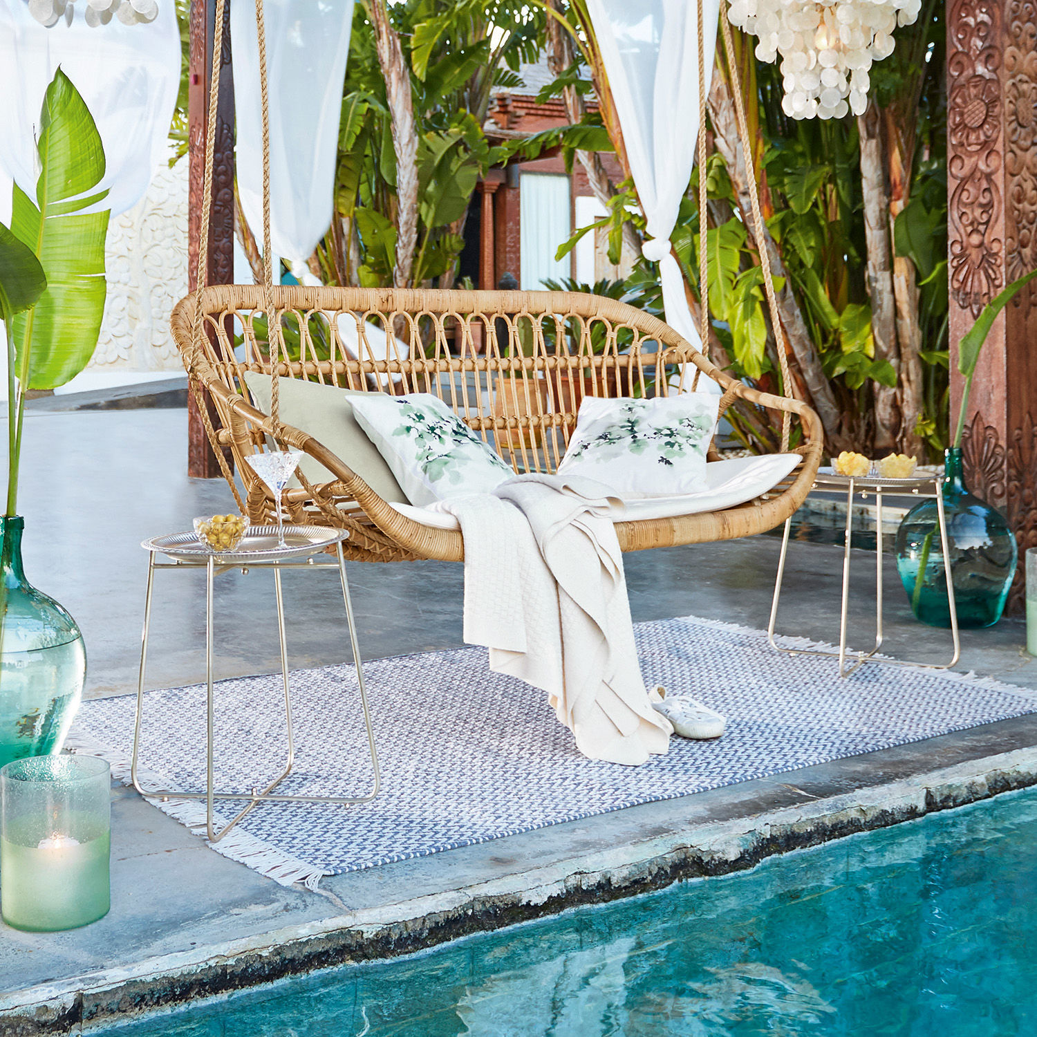 Rattanschaukel in Naturfarbe mit Kissen und Decke am Pool in tropischem Garten