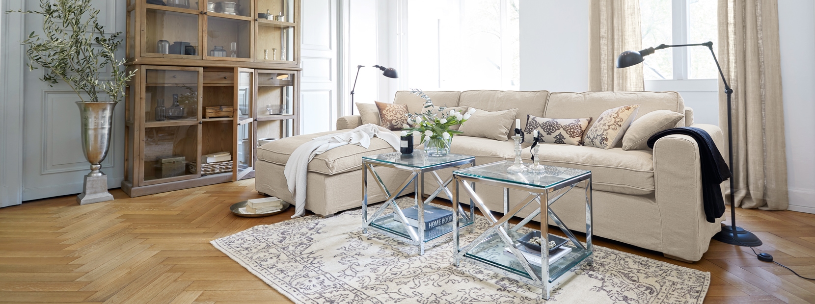 Klassisch elegantes Wohnzimmer mit beigem Sofa, gläsernen Tischen und Holzvitrine auf hellem Parkett