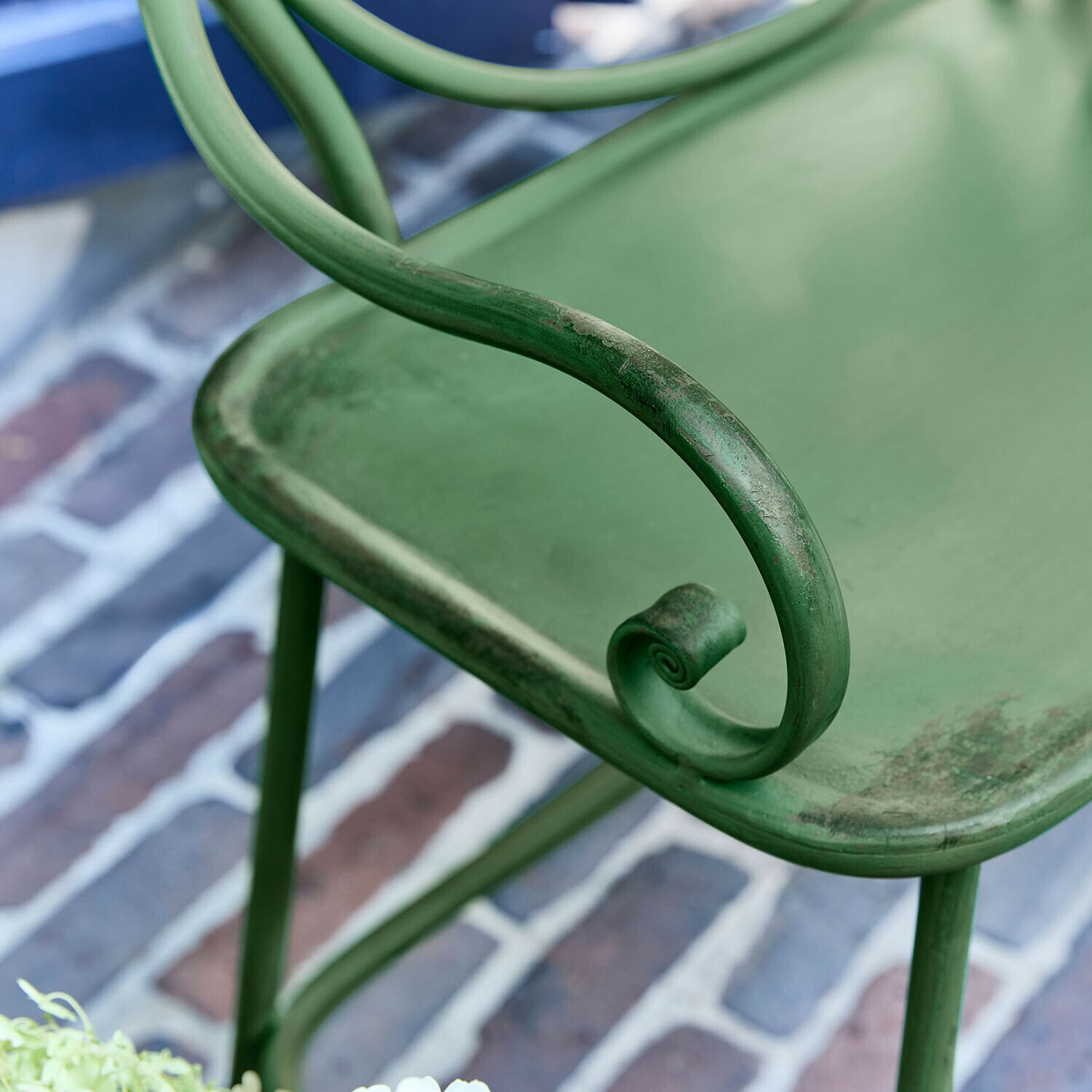 Grüner Metall-Gartenstuhl mit dekorativem geschwungenem Armlehnen-Detail auf Ziegelboden
