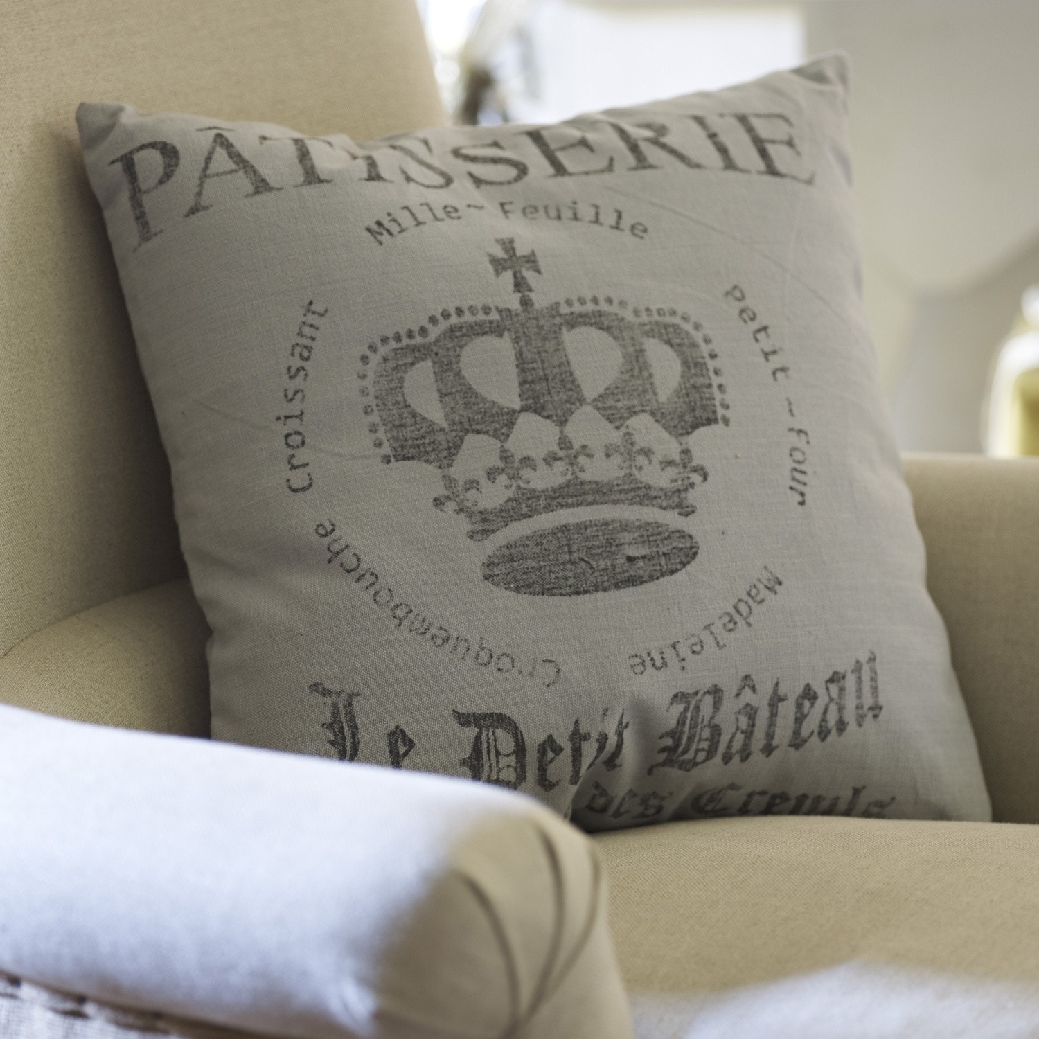 Kissen Ménoire Graues Leinenkissen mit Kronenmotiv auf beigem Sessel als Wohnaccessoire