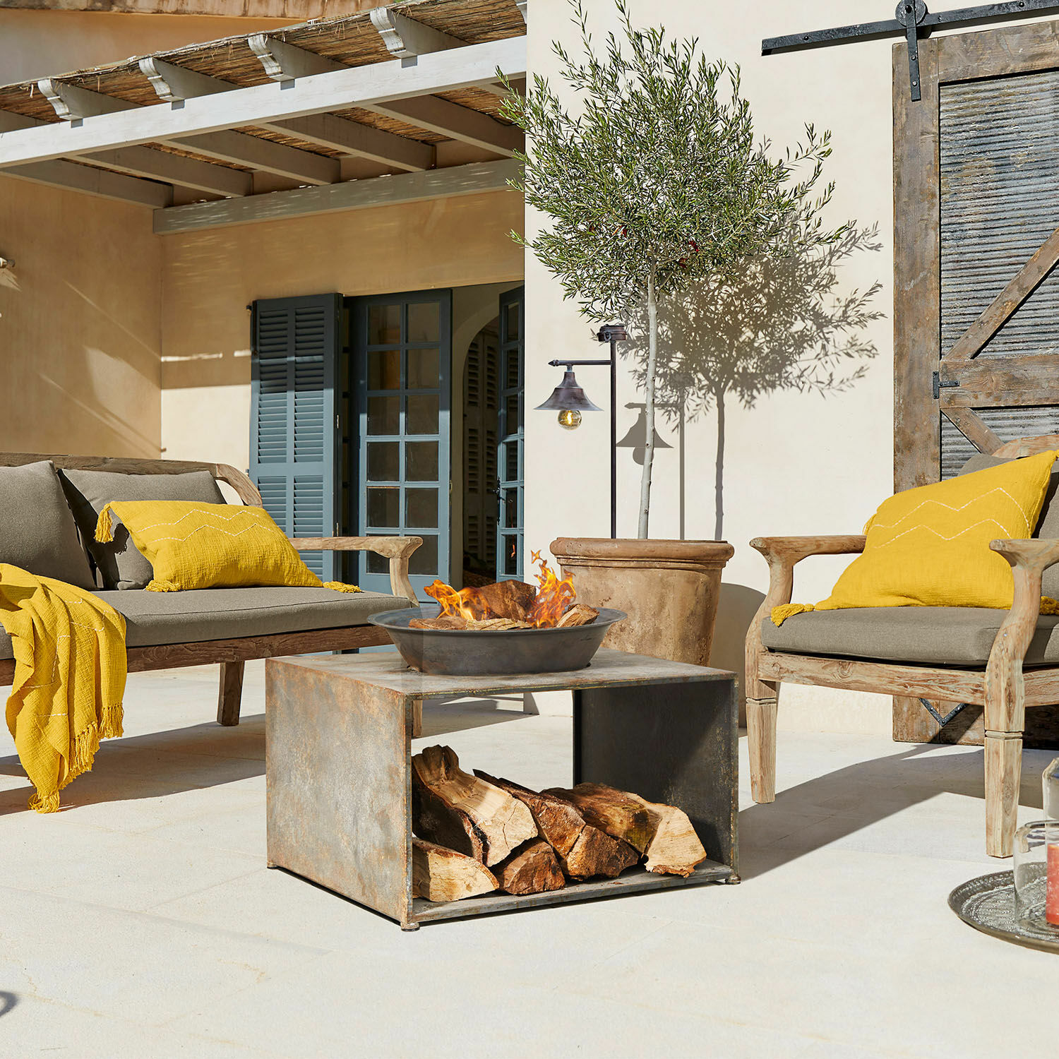 Moderner Metalltisch mit Feuerstelle und Holz auf Terrasse mit Gartenmöbeln und gelben Kissen