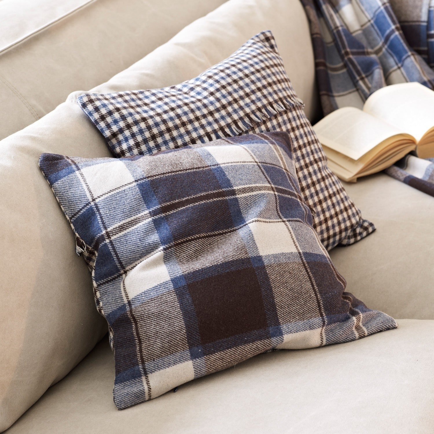 Kissen Clifton jr. Zwei dekorative Kissen mit blau-braunem Karomuster auf beigem Sofa mit offenem Buch