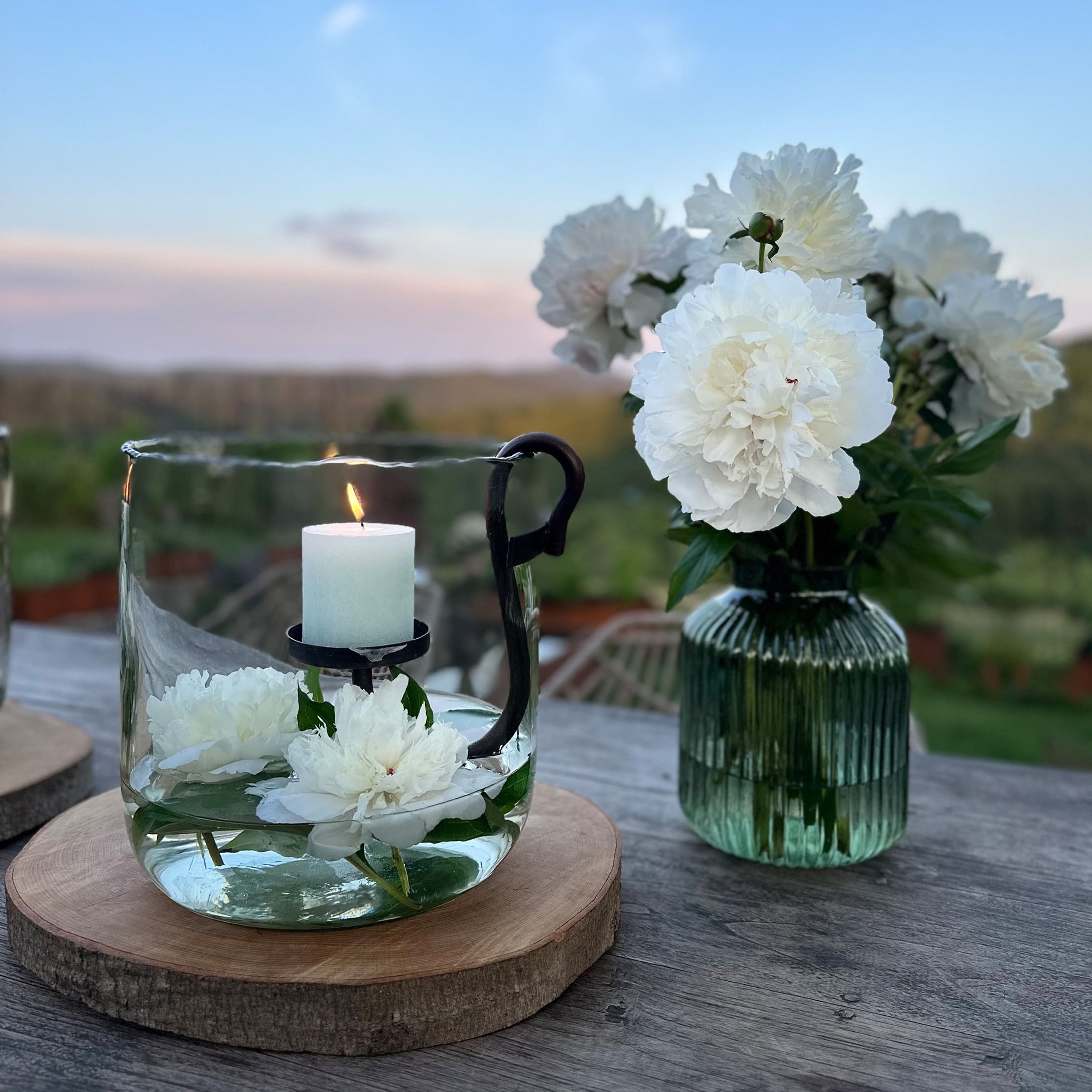 Glaswindlicht mit weißer Kerze und weißen Blumen auf Holzunterlage im Außenbereich