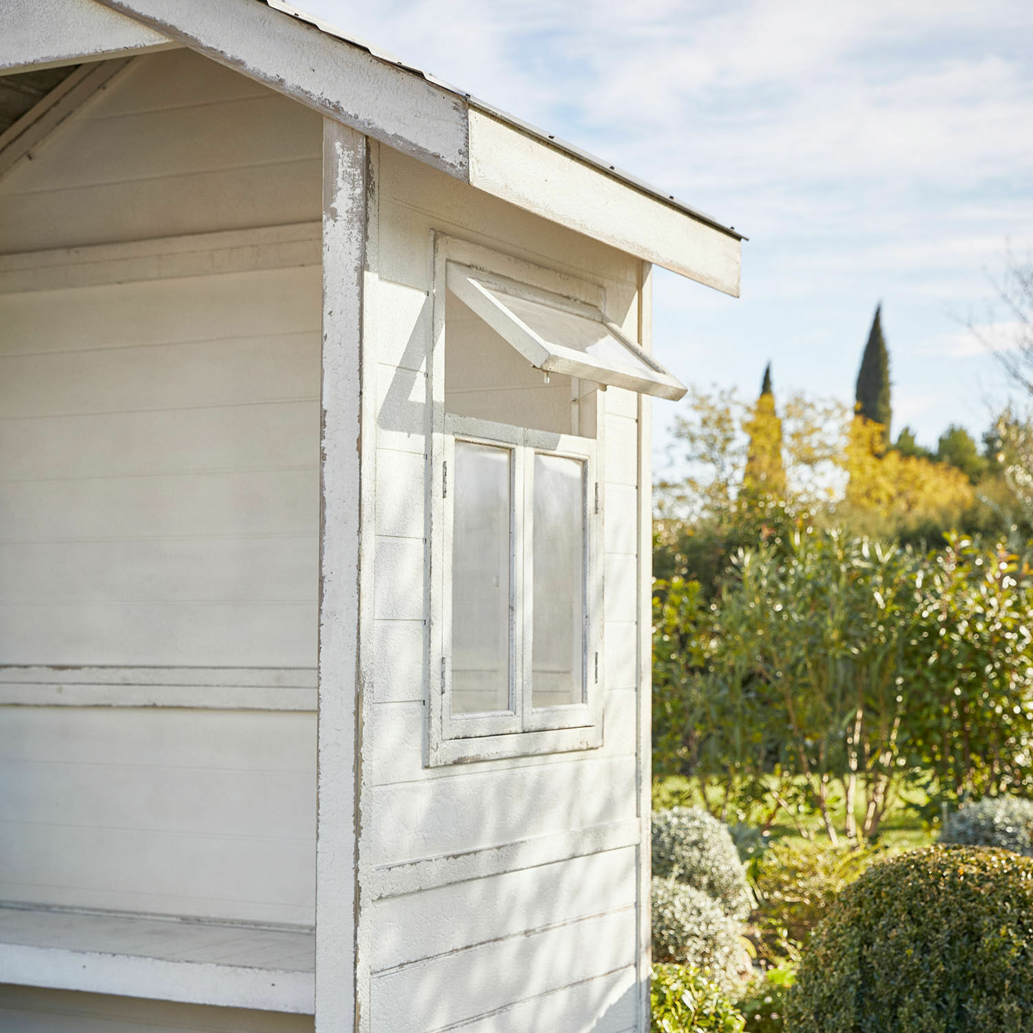 Weißes Gartenhaus aus Holz mit offenem Fenster vor grünem Garten und blauem Himmel