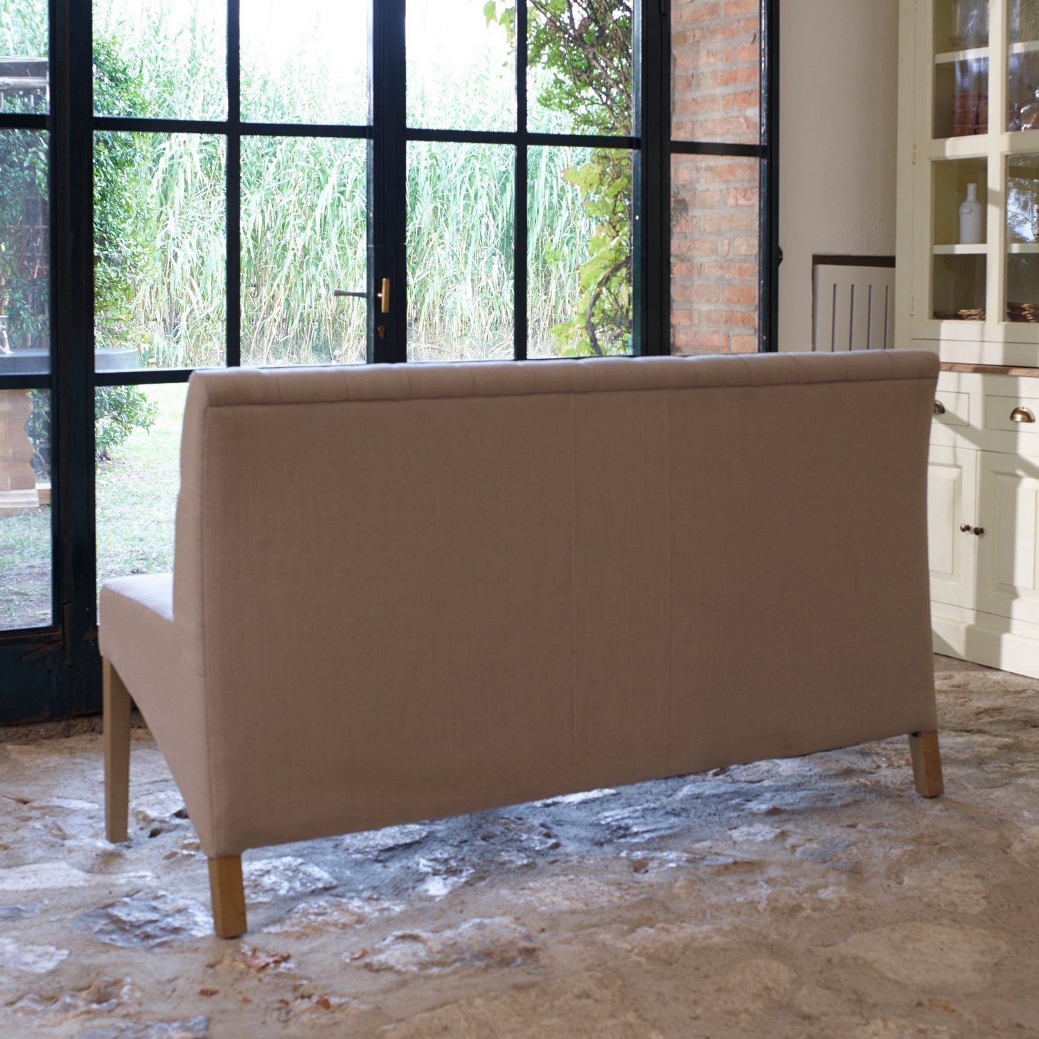 Beige gepolsterte Sofa mit Holzbeinen auf Steinboden vor großem Fenster