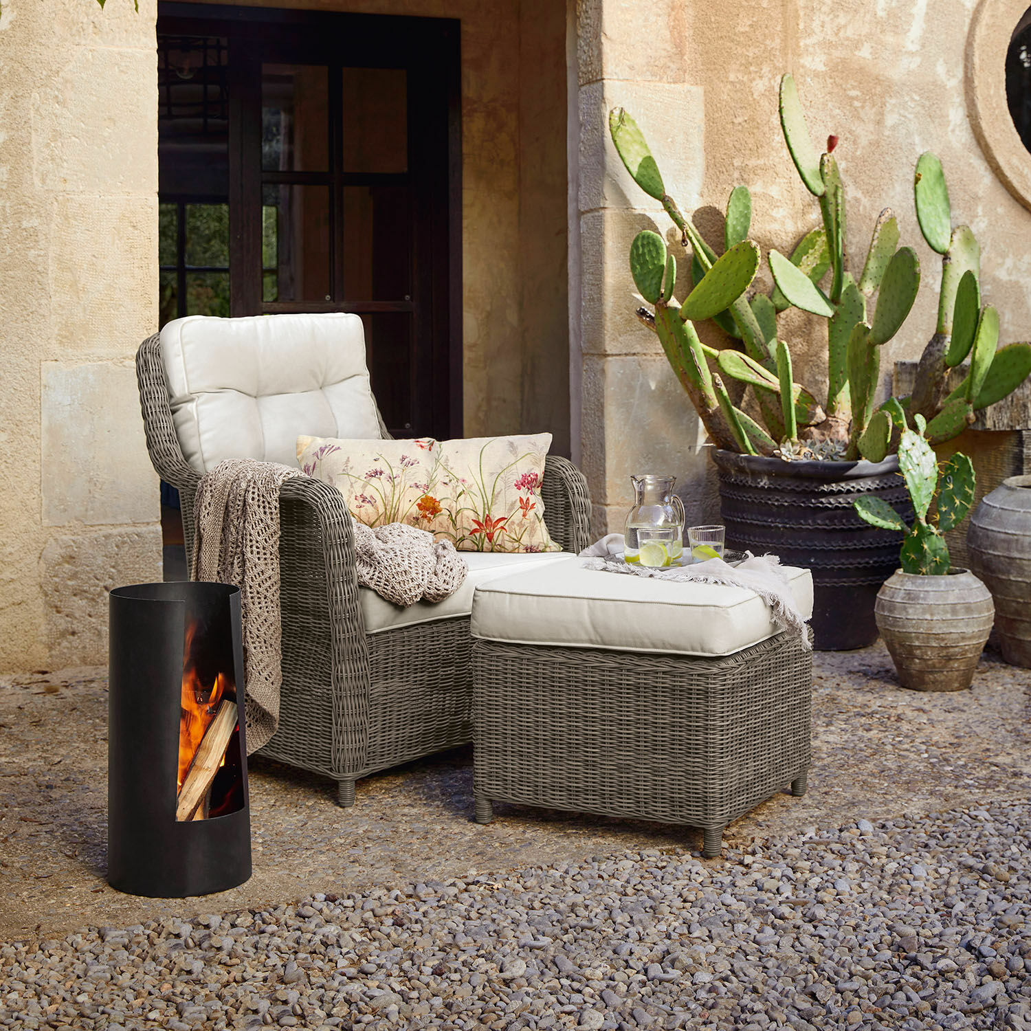 Grauer Rattan-Sessel mit cremefarbenem Polster und Hocker auf Terrasse mit Kakteen und Feuerstelle