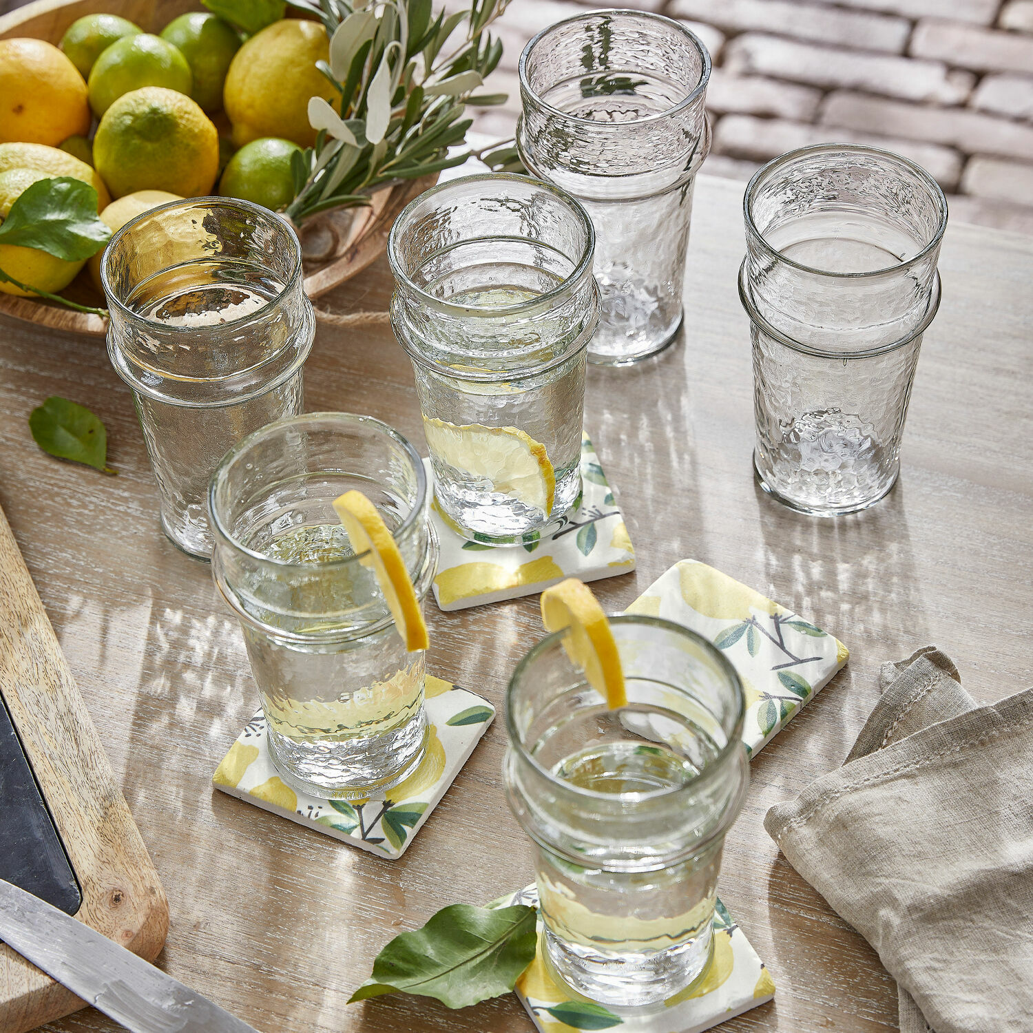 Klarglas-Gläser mit Struktur, teils mit Zitronenscheiben, auf Holztisch mit Zitrusfrüchten