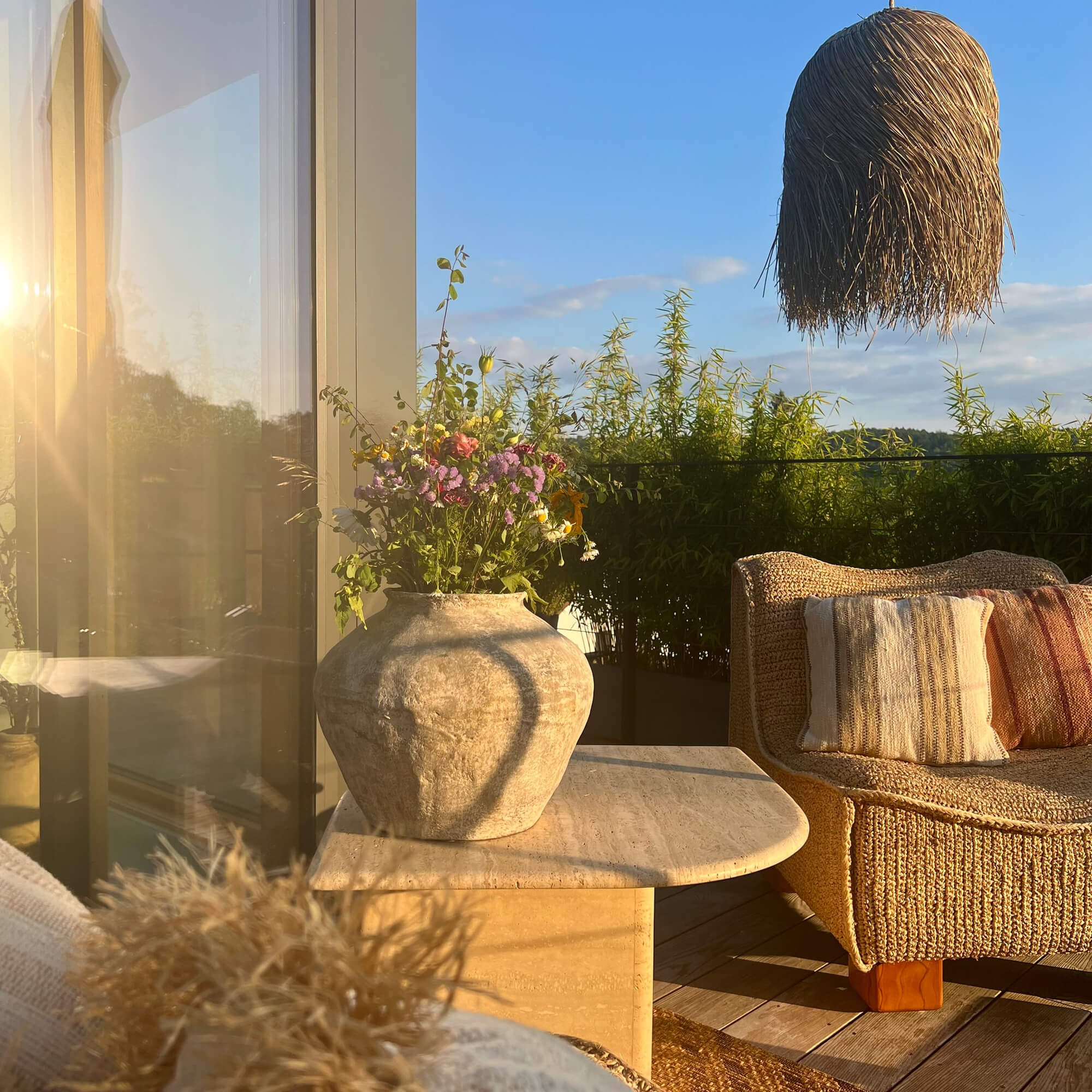 Große Steinvase auf Tisch neben Korbsofa mit Kissen auf begrüntem Balkon bei Sonnenlicht