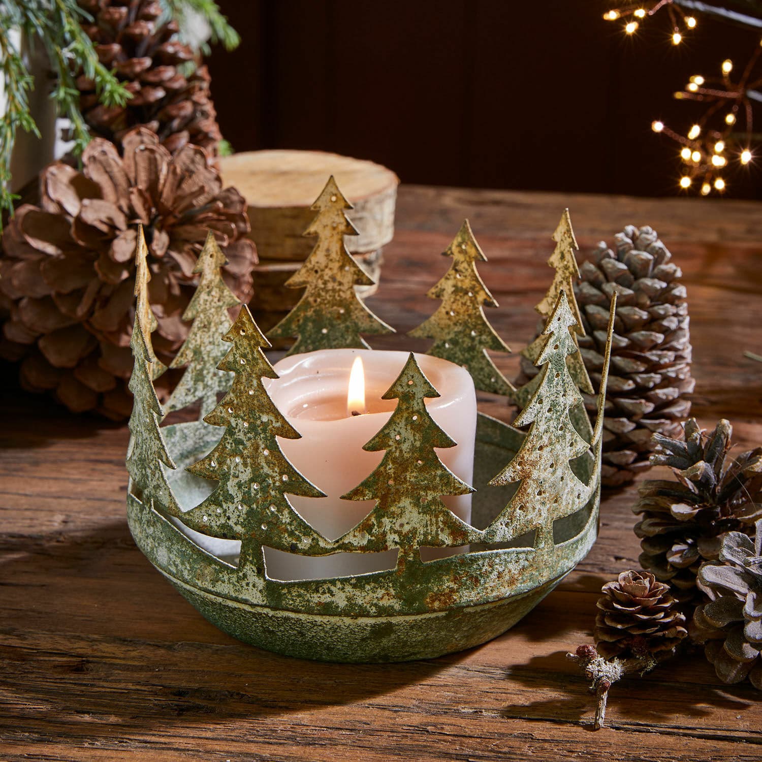 Windlicht Norundra Metall-Kerzenhalter mit Tannenbaum-Motiv in grünem Used-Look auf Holztisch mit Kiefernzapfen