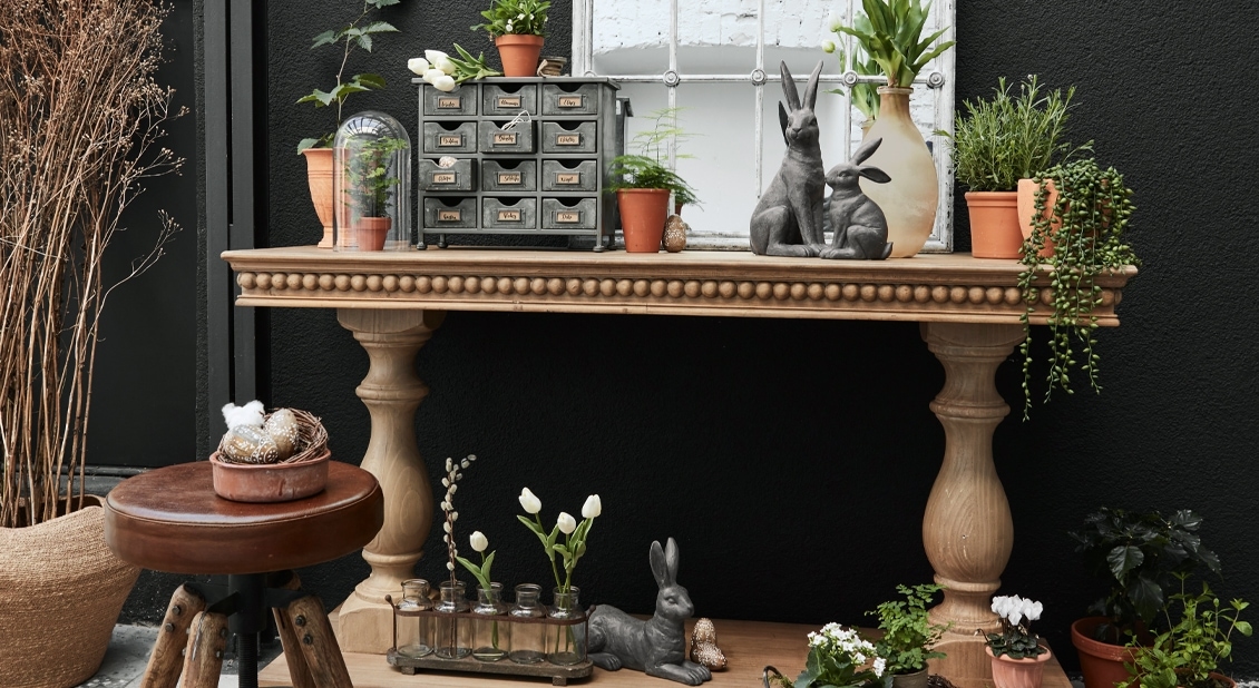 Holztisch mit Hasenfiguren und grünen Pflanzen in Töpfen vor schwarzem Hintergrund dekoriert