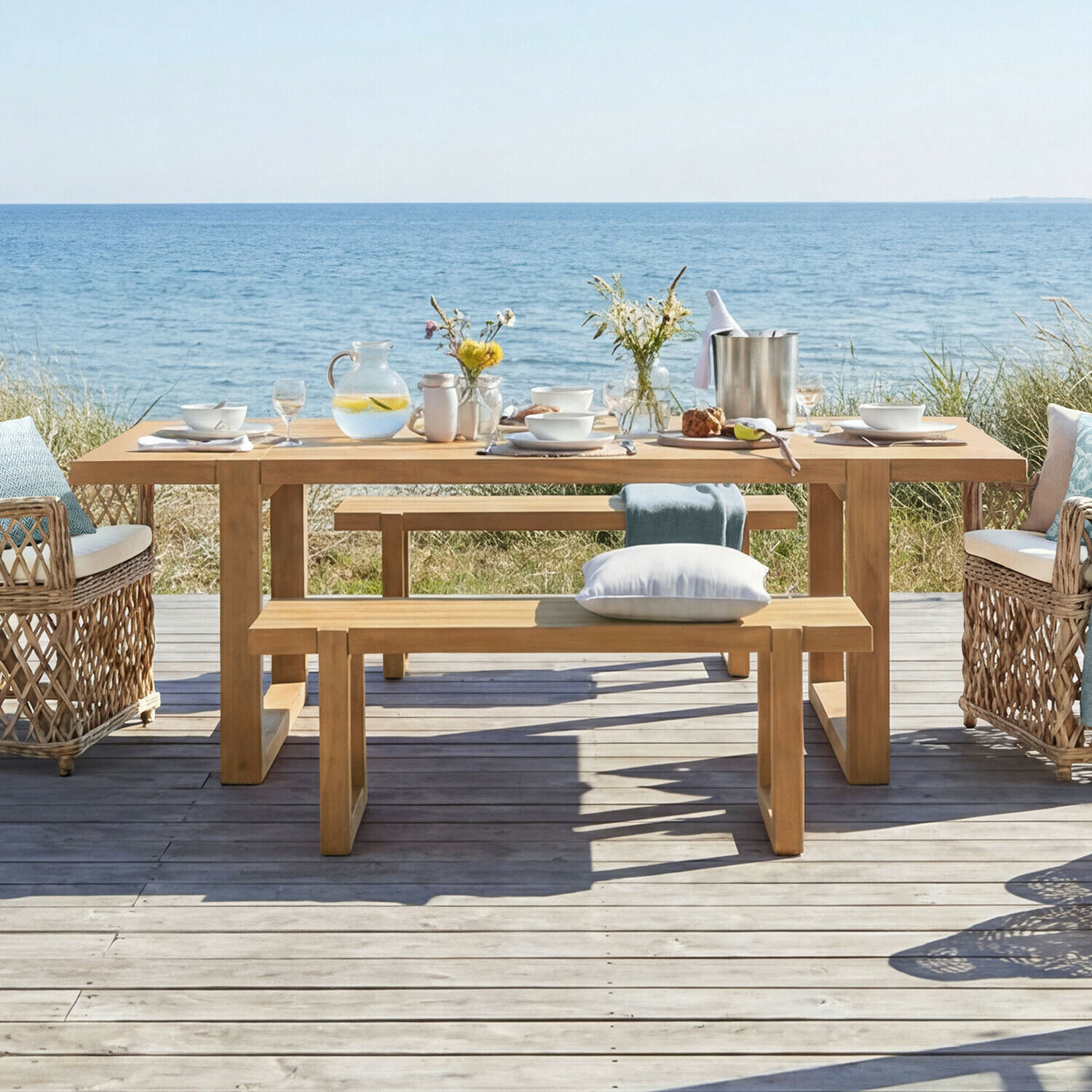 Tisch Halinor Gartentisch aus Holz mit Bänken und Korbsesseln am Meer, dekoriert mit Geschirr und Blumen