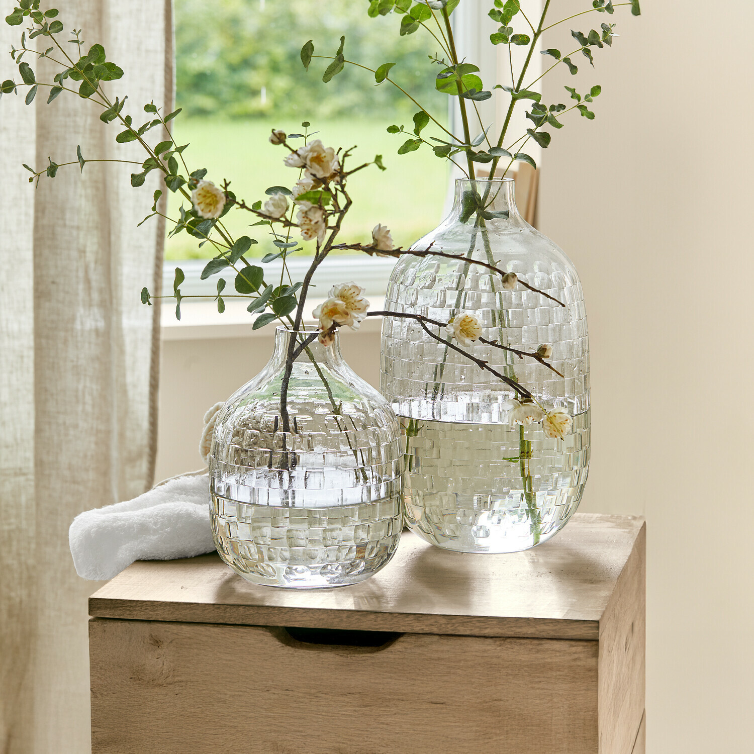 Vase 2er Set Qervilon Zwei durchsichtige Glasvasen mit Karomuster und grünen Zweigen mit Blüten auf Holztisch