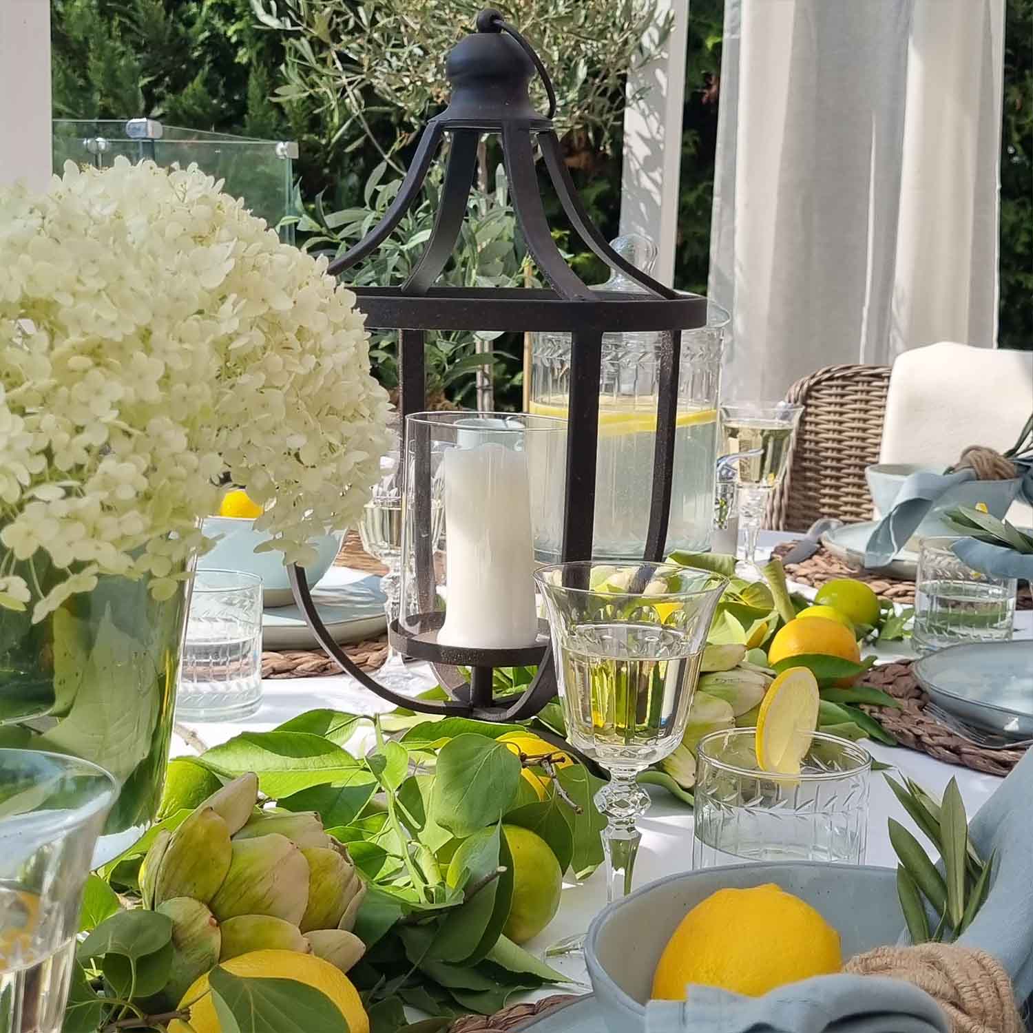 Schwarzer Metall-Lampion mit weißer Kerze auf sommerlich dekoriertem Tisch mit Zitronen und grünen Blättern