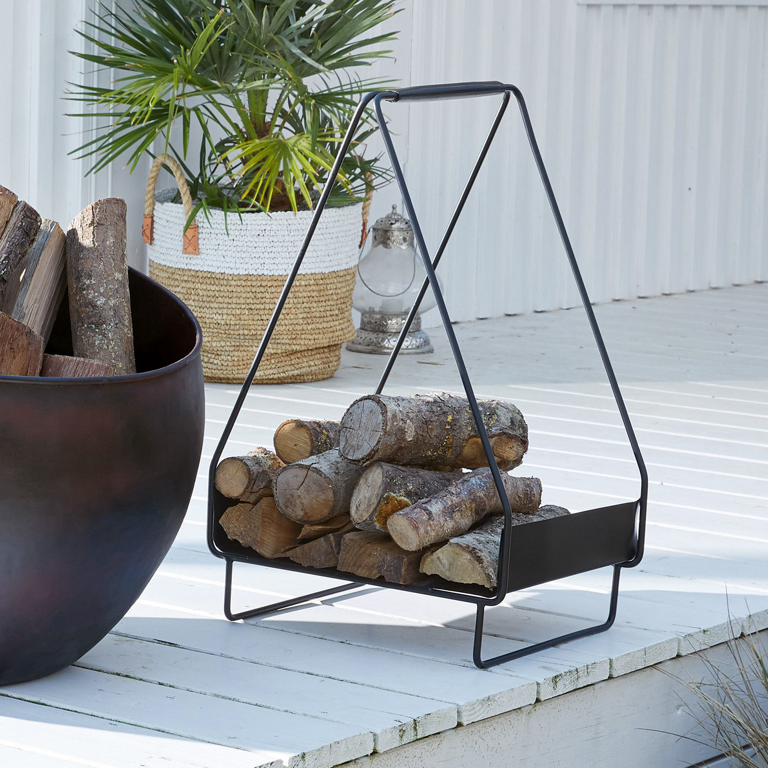 Schwarzer Metall-Holzkorb mit Holzscheiten auf weißem Terrassenboden im Außenbereich