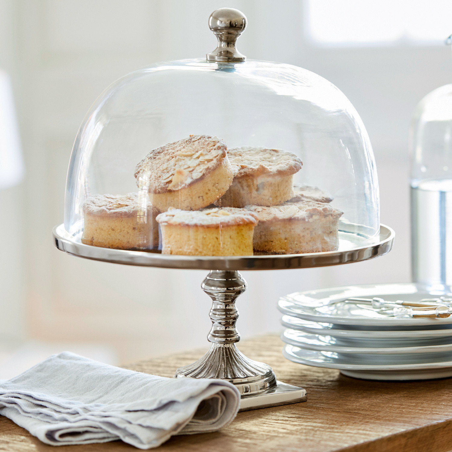 Glasglocke mit Platte Mine Metall-Tortenplatte mit transparenter Glasglocke und Kuchen auf Holztisch neben weißen Tellern