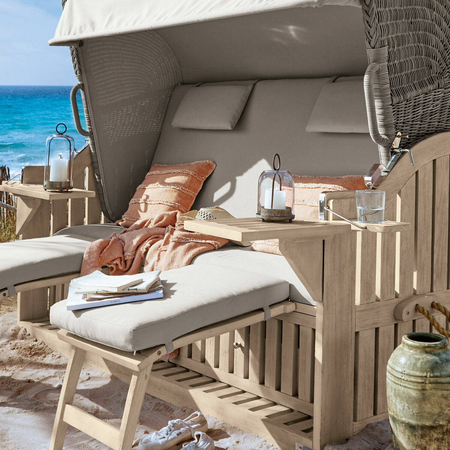 Strandkorb aus Holz mit hellgrauen Polstern und orangefarbener Decke am Meer