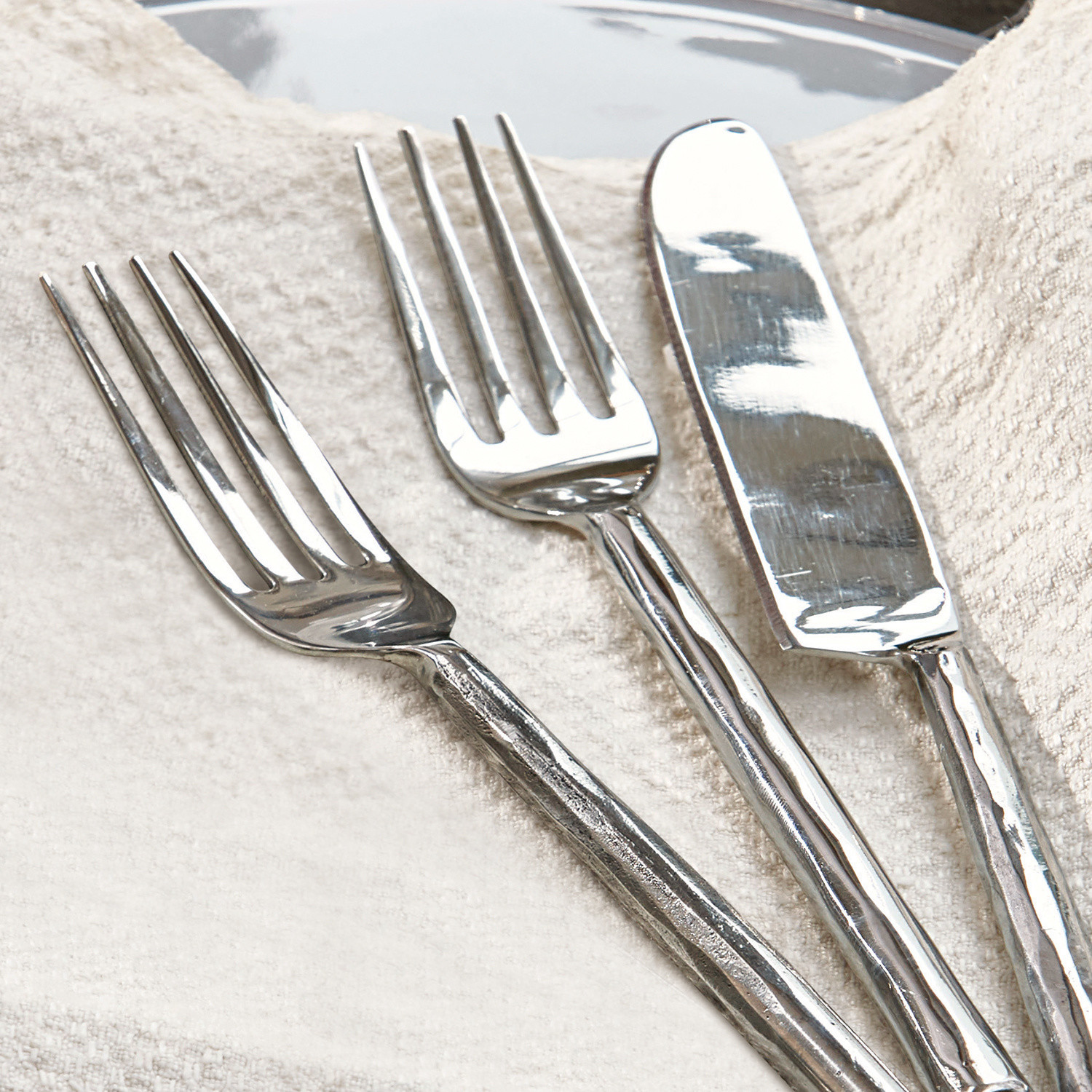 Silbernes Besteck mit unregelmäßiger Struktur an den Griffen auf hellem Stoff, elegantes Tischset