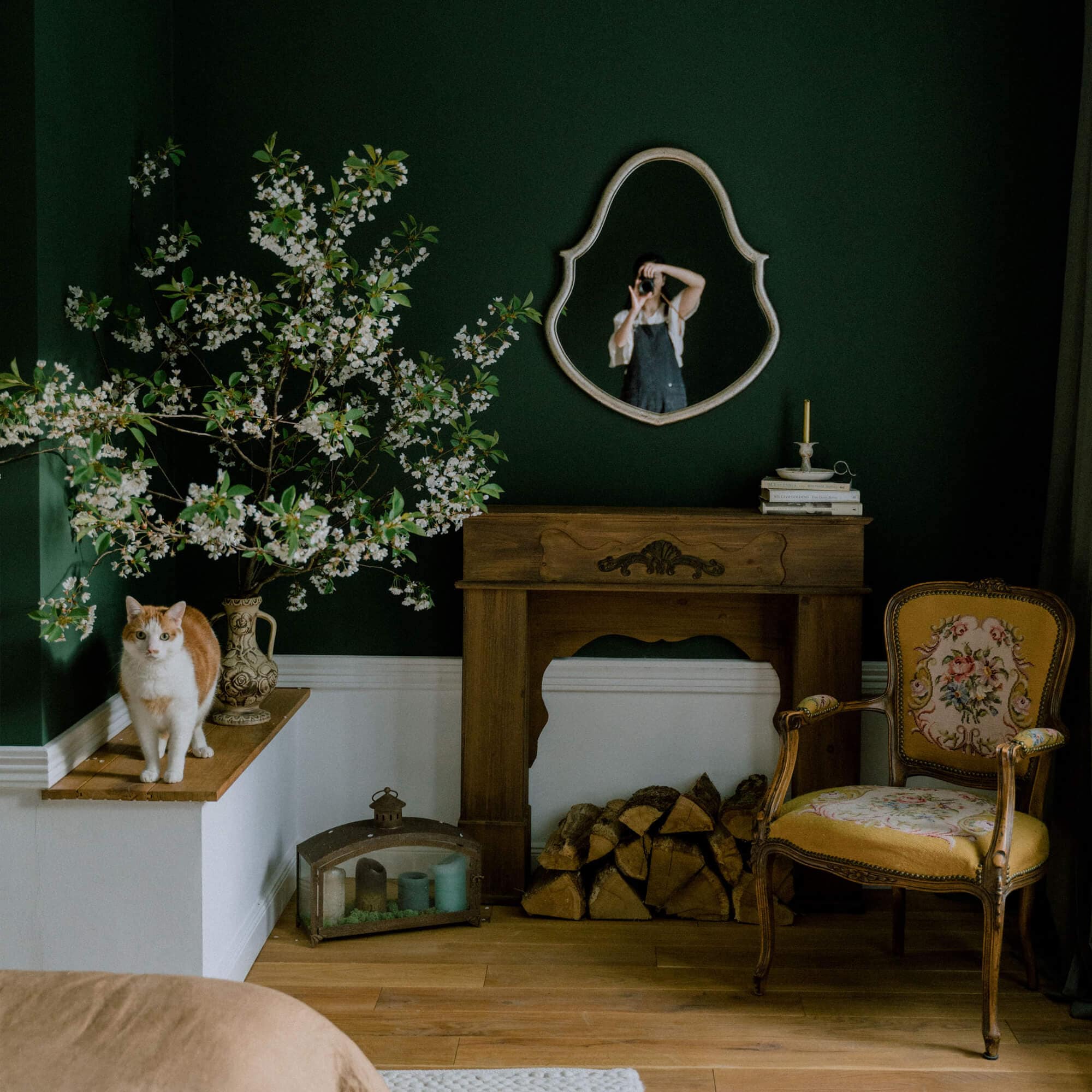 Gemütliches Wohnzimmer mit dunkelgrüner Wand, Holz-Kamin, gelbem Blumenstuhl und Katze auf der Ablage