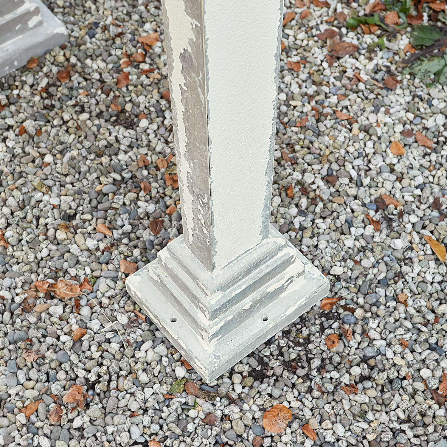 Postament aus cremefarbenem, antik wirkendem Holz auf Kies mit Laub als Gartendekoration