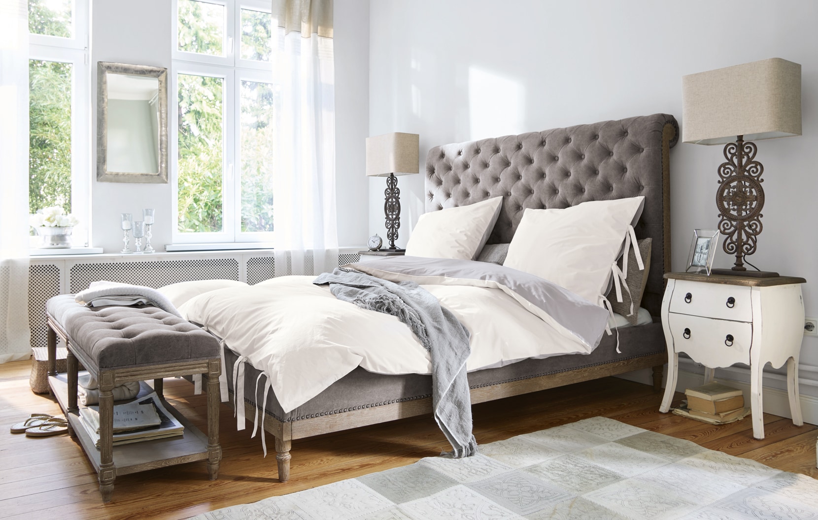 Klassisches Schlafzimmer mit grauem Polsterbett und weißer Bettwäsche in hellem Raum