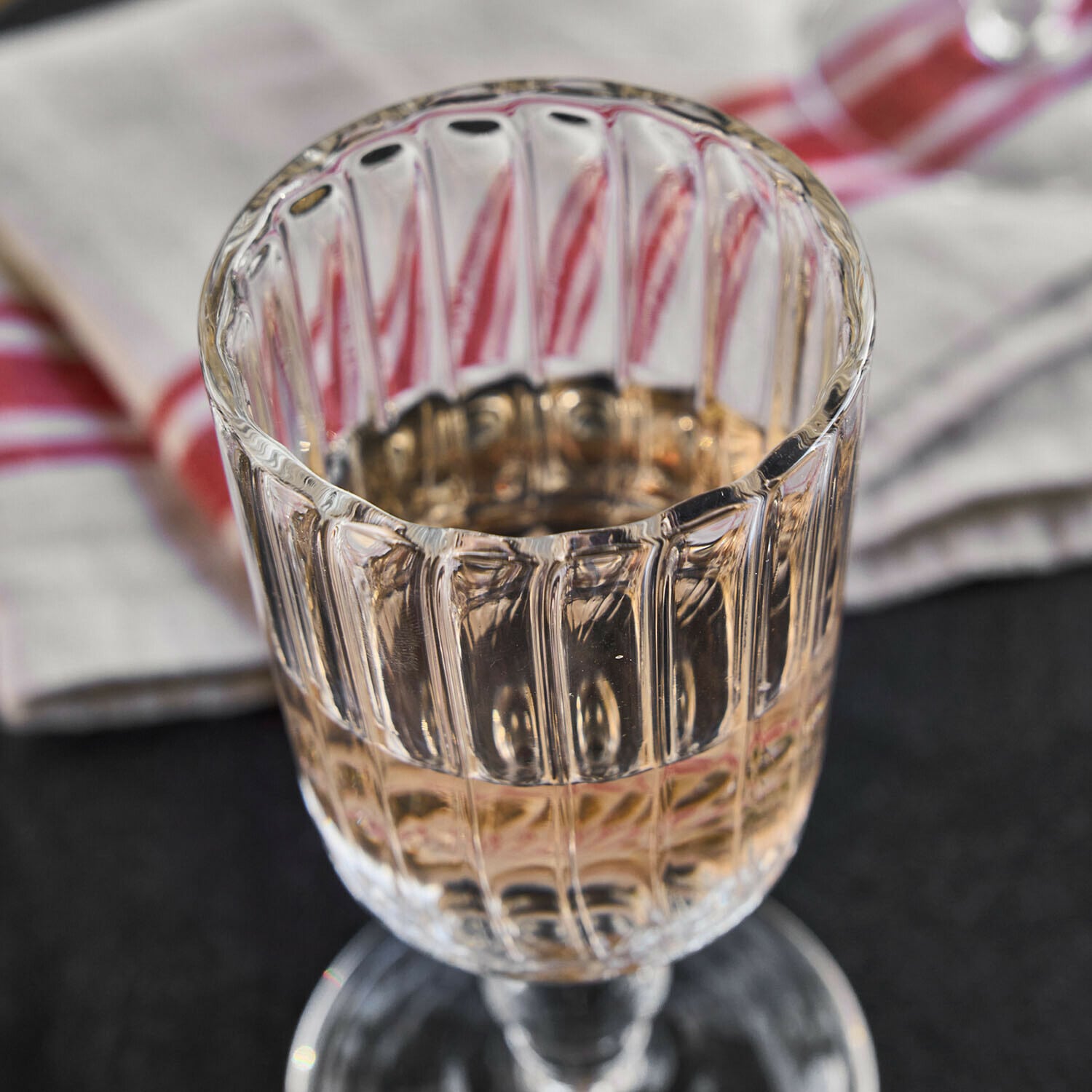 Klarer, gerillter Glas-Weinkelch mit hellem Getränk auf Tisch mit Stoff im Hintergrund